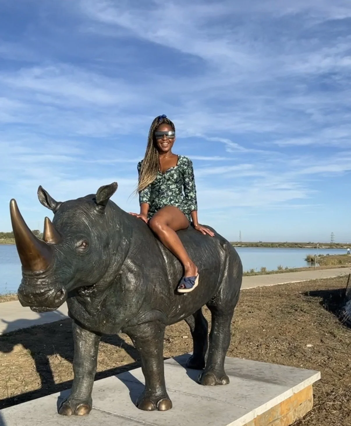 A woman with long braids and sunglasses sits on a large rhinoceros sculpture outside near water under a blue sky with clouds.