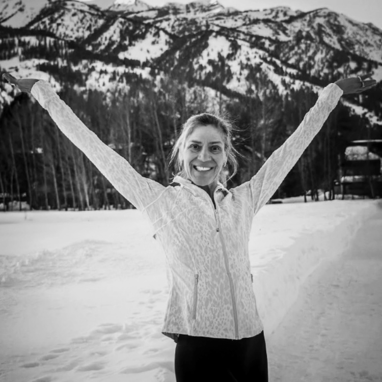 A woman standing outdoors in a snowy landscape with mountains in the background, smiling with her arms raised.