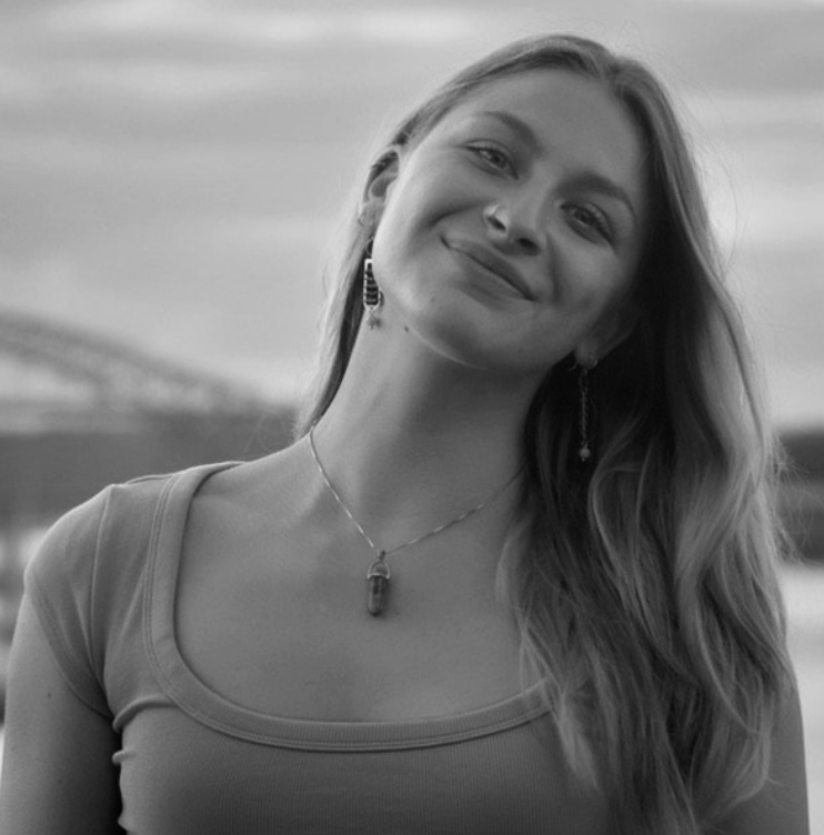 A young woman with long hair smiling outdoors, wearing earrings, a necklace, and a T-shirt, with a blurred landscape background.