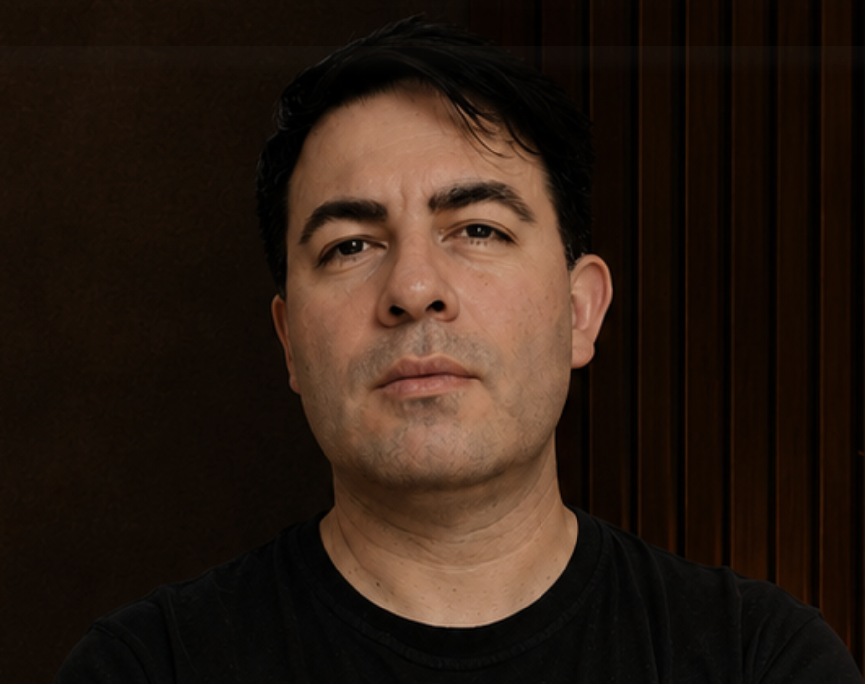 Portrait of a man with dark hair, wearing a black shirt, standing against a brown wooden background.