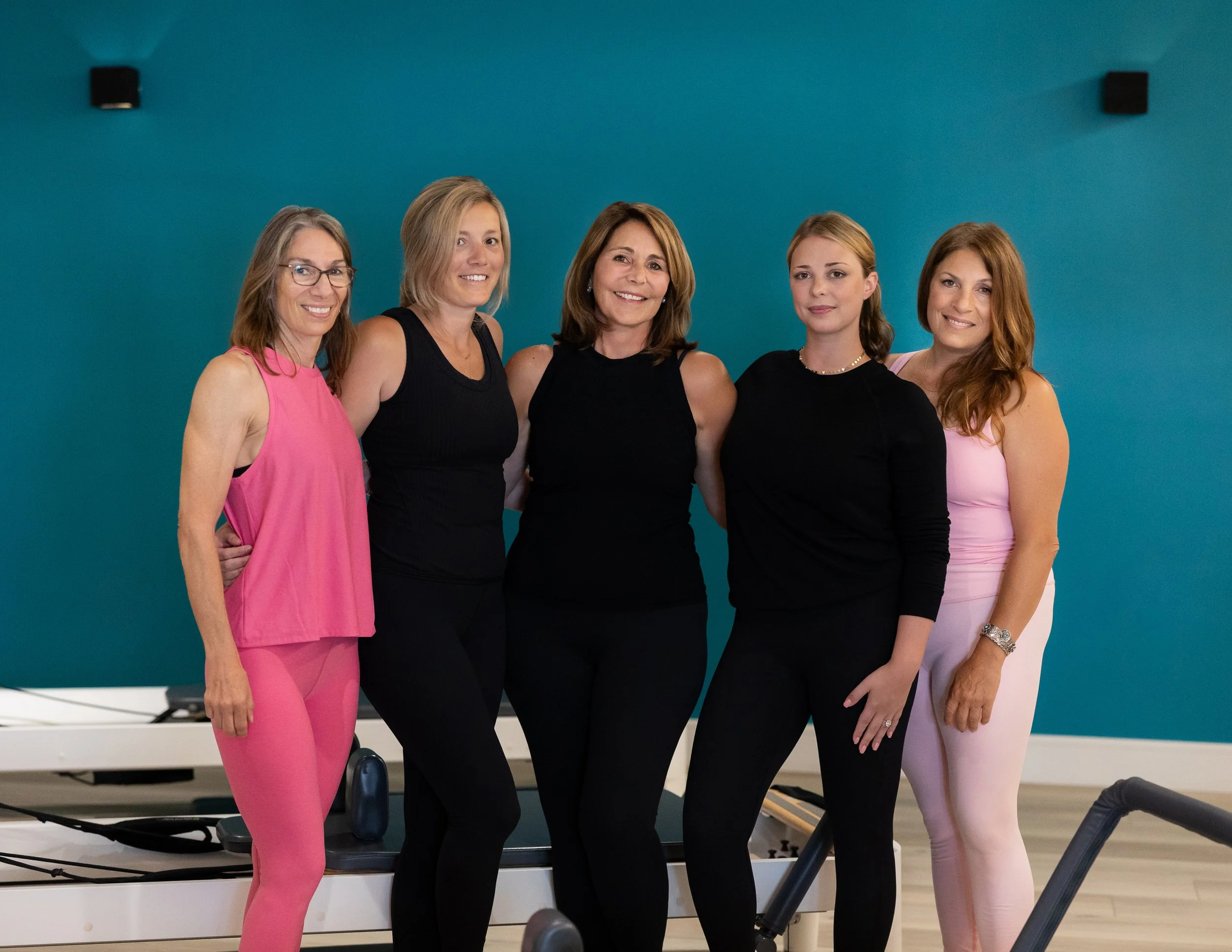 Five women with medium-length hair, wearing sleeveless or fitted tops, standing together in a room with a blue wall and fitness equipment, smiling at the camera.