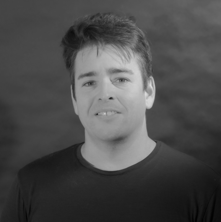 Black and white portrait of a man with short, tousled hair, wearing a dark T-shirt, smiling slightly, against a blurred background.