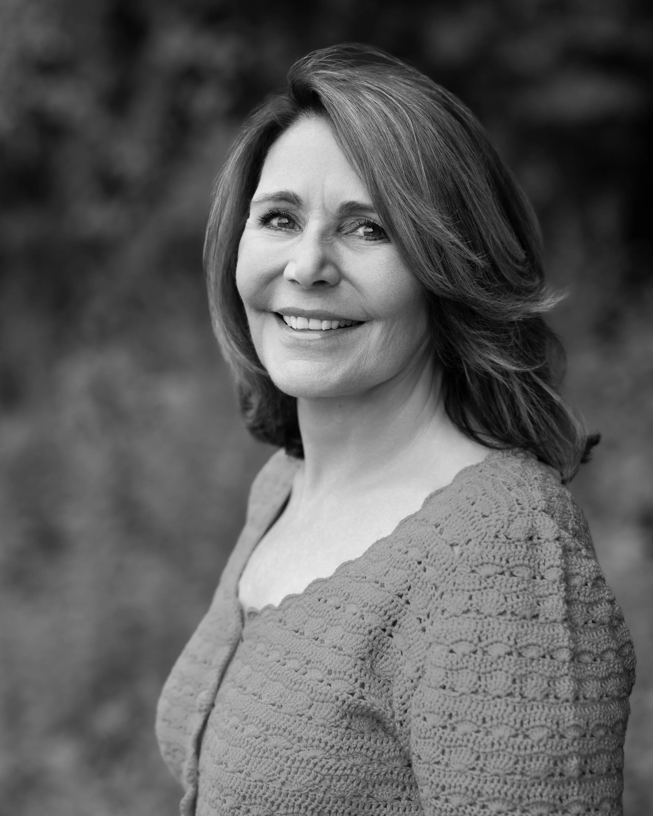 Black and white portrait of a smiling woman with shoulder-length hair, wearing a textured sweater, outdoors in a natural setting.