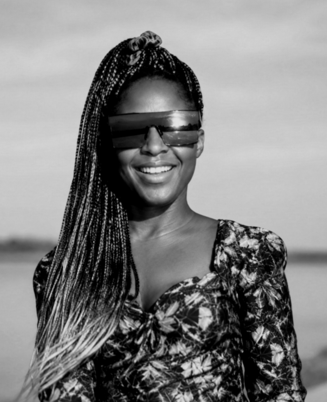 A smiling woman with braided hair and sunglasses standing outdoors, with a blurred background.