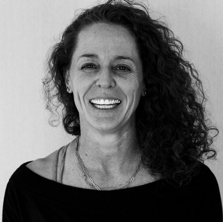 A black and white portrait of a woman smiling with curly hair, wearing earrings, layered necklaces, and a dark top.