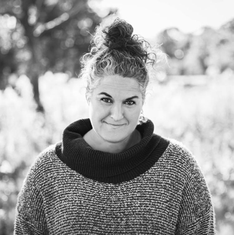 A woman with curly hair tied in a bun, wearing a textured sweater with a large, turtleneck collar, standing outdoors with blurred trees in the background.