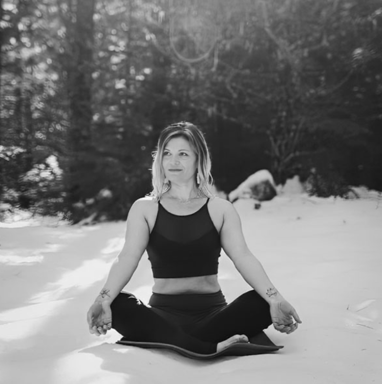 A woman practicing yoga in a forested area with snow, sitting cross-legged on a mat and looking peaceful.