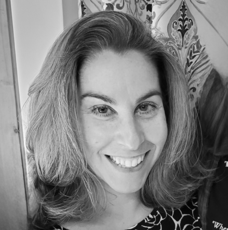 Black and white portrait of a smiling woman with shoulder-length hair, indoors with patterned curtains or wallpaper in the background.