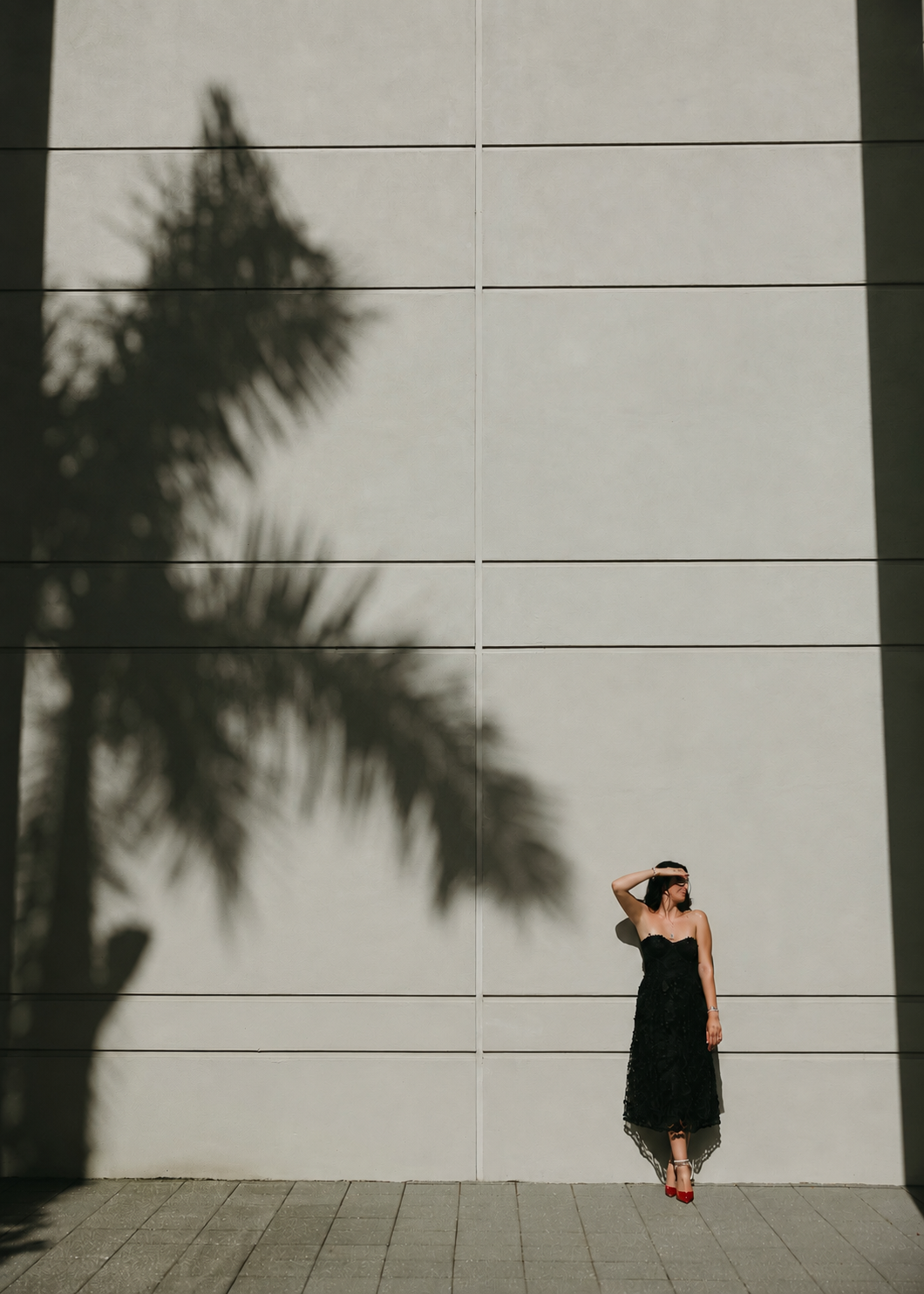 A woman in a black dress and red heels stands against a light-colored tiled wall, shielding her eyes from the sun with her right hand. Her shadow and the shadow of a palm tree are cast on the wall.