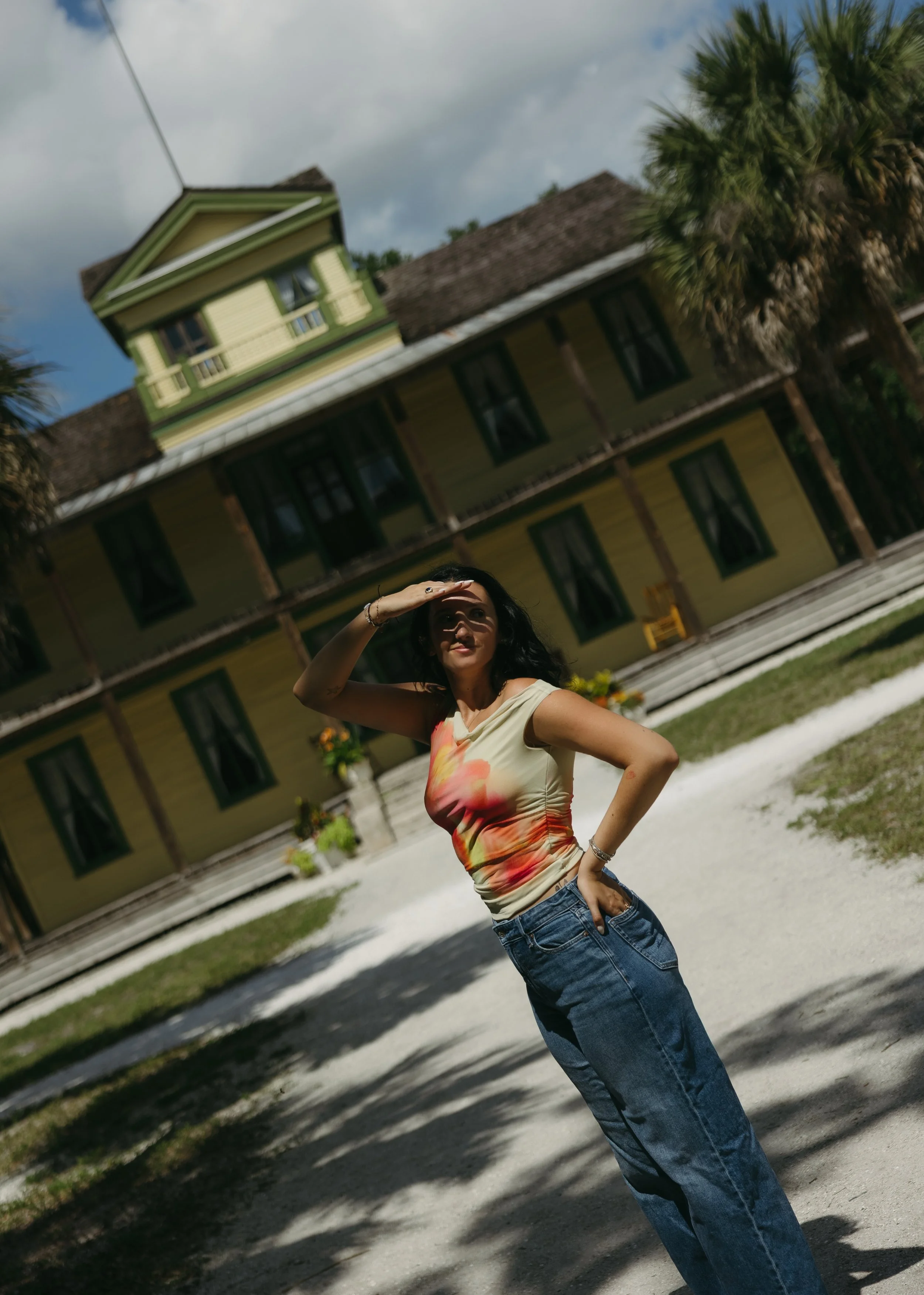 A woman with dark hair wearing a colorful sleeveless top and jeans, standing outdoors in front of a yellow house with a green trim and palm trees, shading her eyes from the sun.