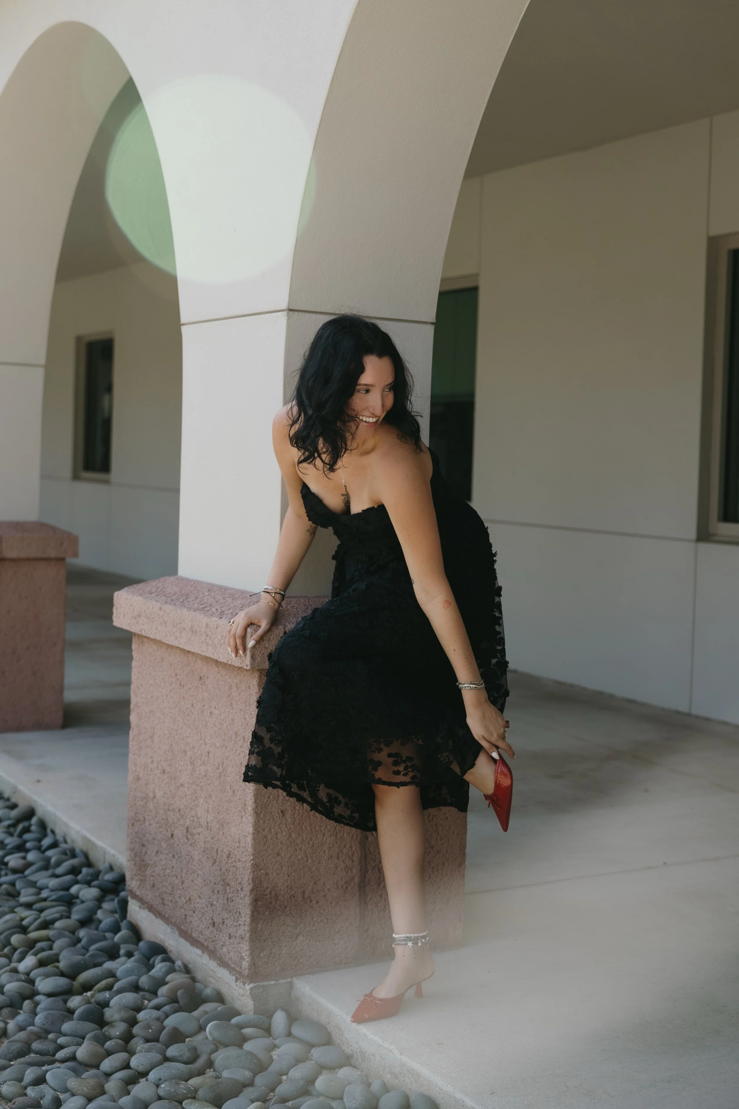 A woman in a black lace dress and red heels sitting on a pink concrete ledge outdoors, smiling and looking to her right.