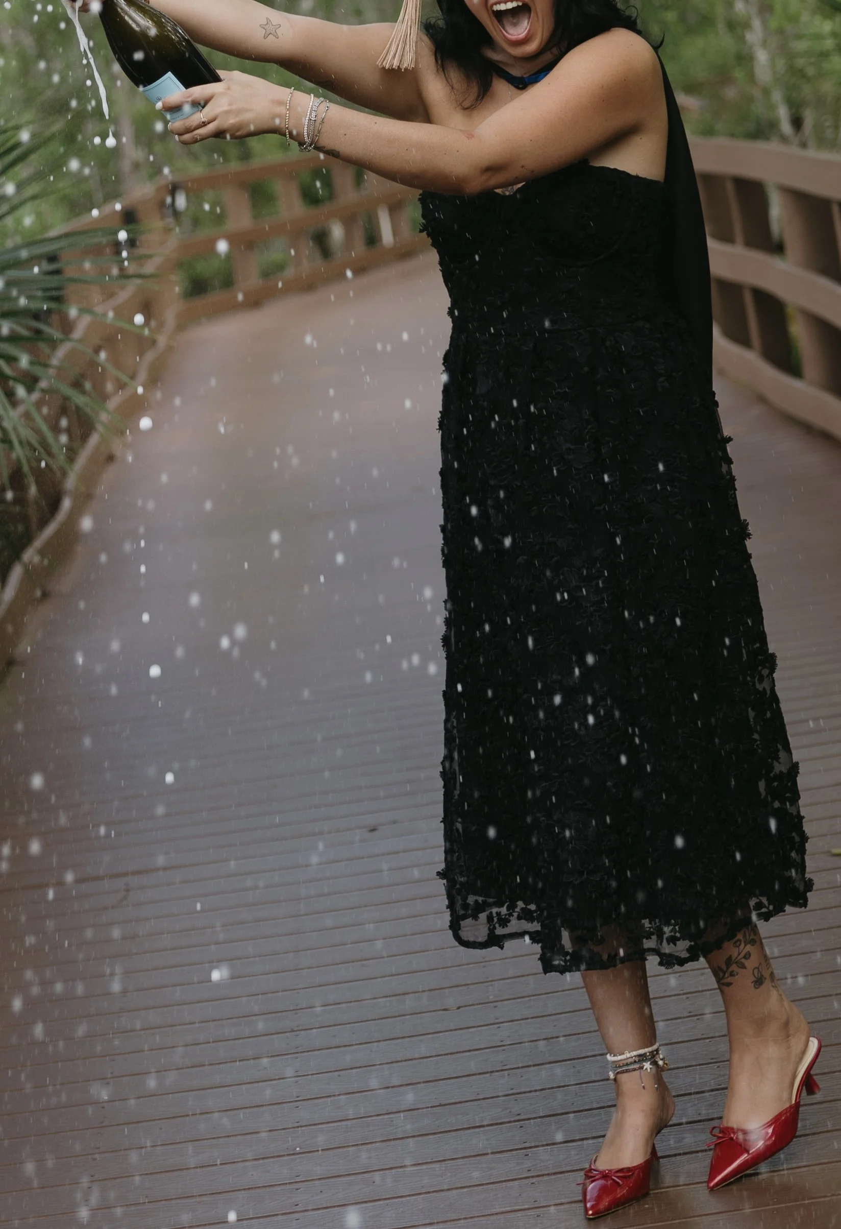A woman in a black lace dress and red high heels is celebrating on a wet wooden deck, spraying champagne. She has tattoos on her legs, bracelets on her wrist, and a star tattoo on her arm. The background is blurred greenery.