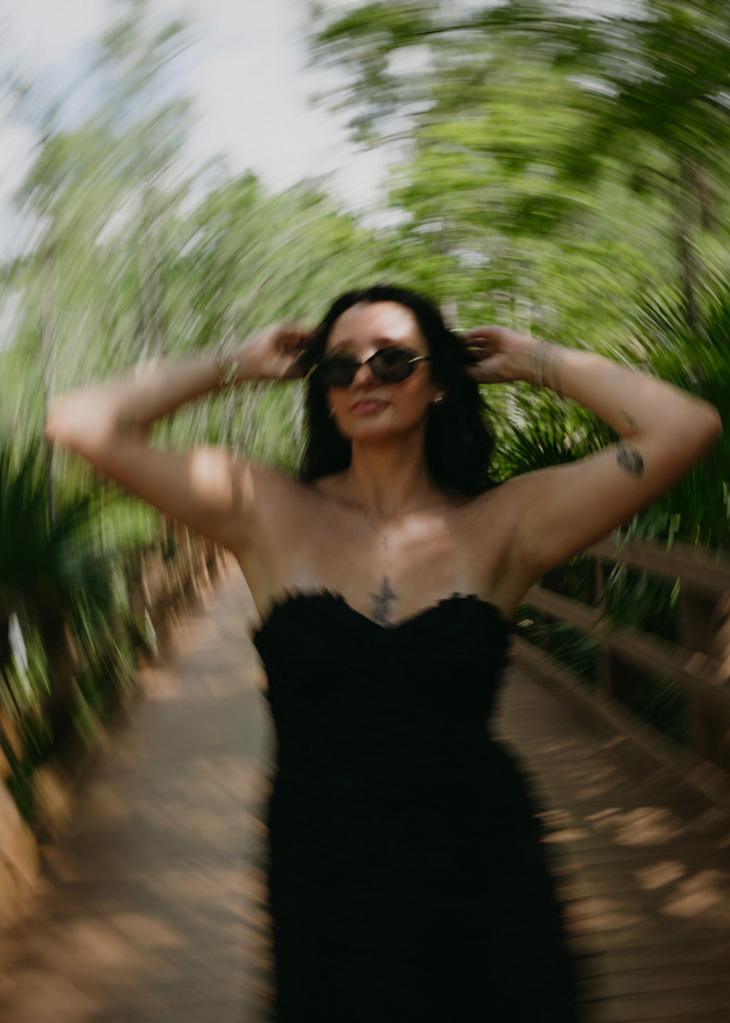 A woman with black hair wearing sunglasses and a strapless black dress standing on a wooden pathway surrounded by greenery, with a motion blur effect.