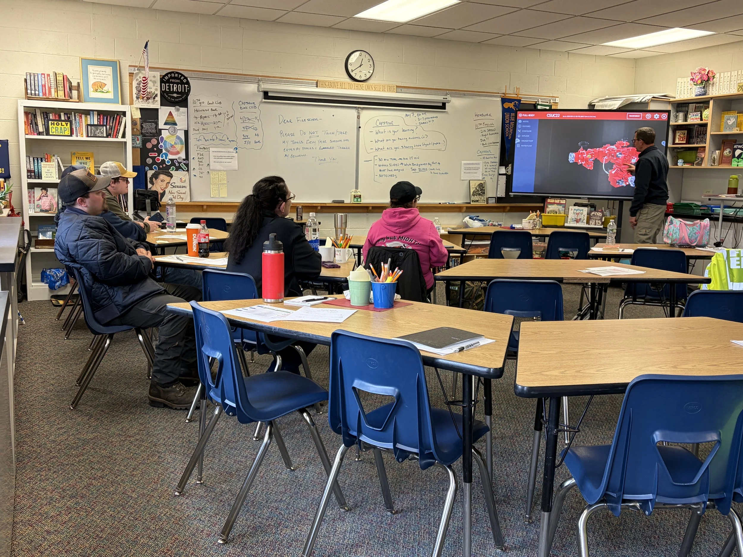 Classroom scene with students sitting at desks facing a teacher and a large digital display. The room has a whiteboard, bookshelves, and decorative items.
