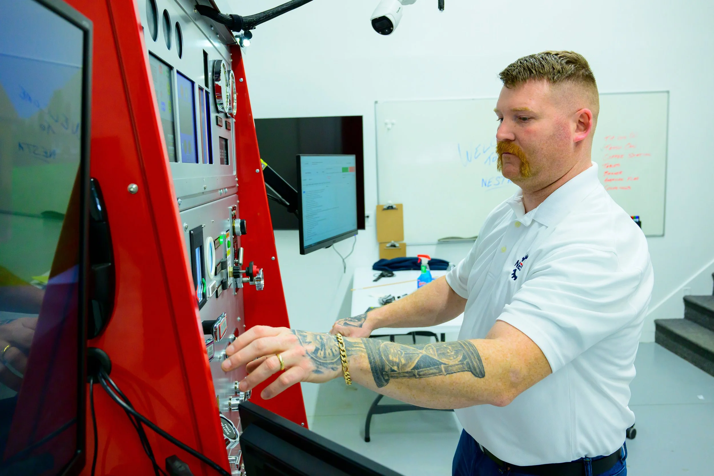 A man working on a red electrical control panel with a gray monitor in the background at a workshop or classroom.