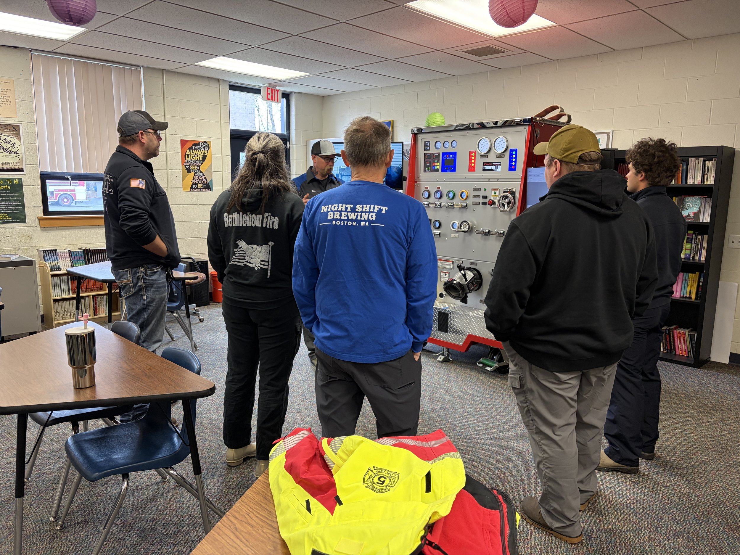 A group of people gathered around a fire truck-themed control panel in an indoor room, with someone explaining or demonstrating something. There are chairs, a bookshelf, posters on the wall, and personal protective gear on the table in the foreground.