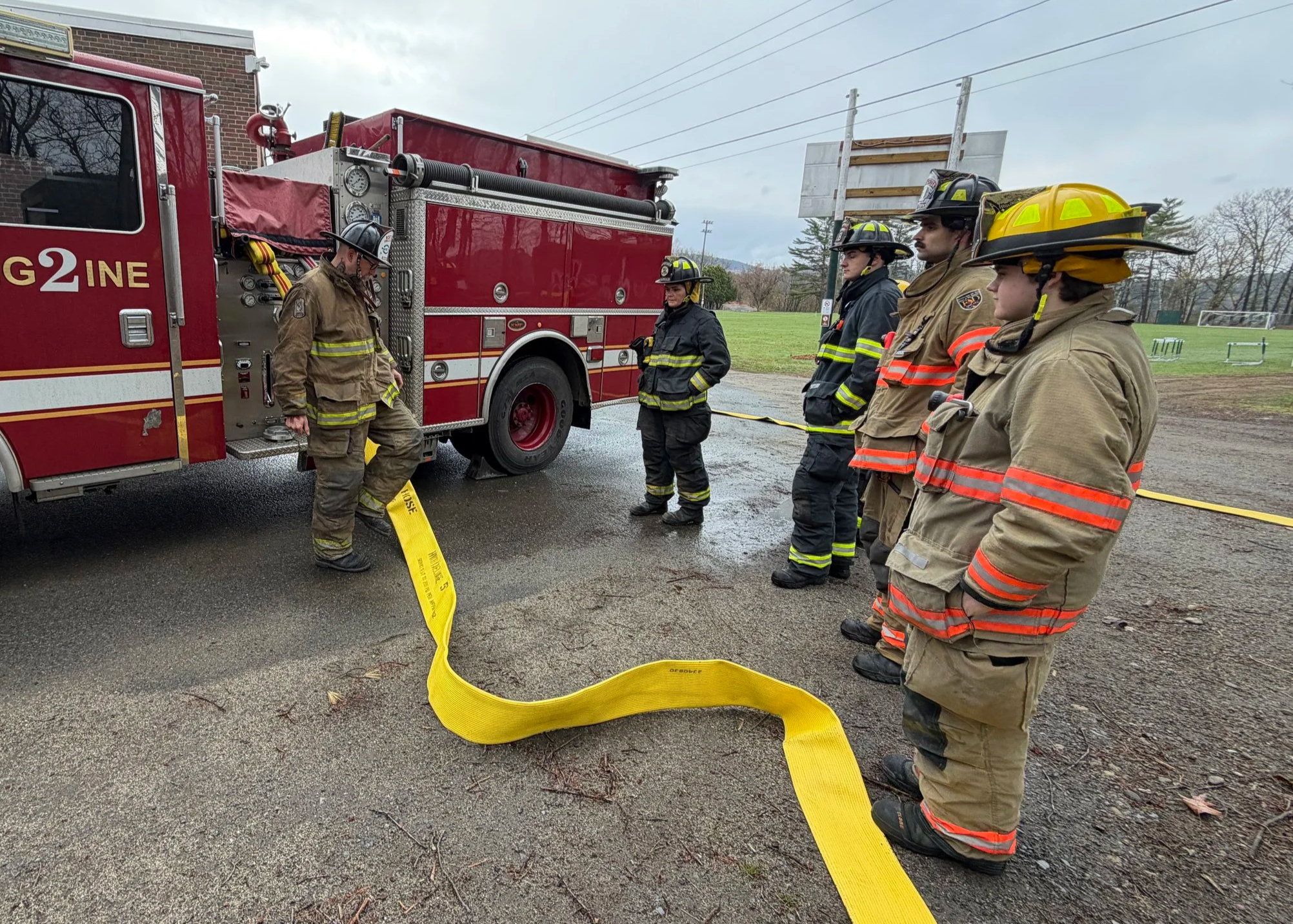 A group of firefighters in protective gear standing beside a red fire truck on a wet street, with a yellow fire hose laid out on the ground.