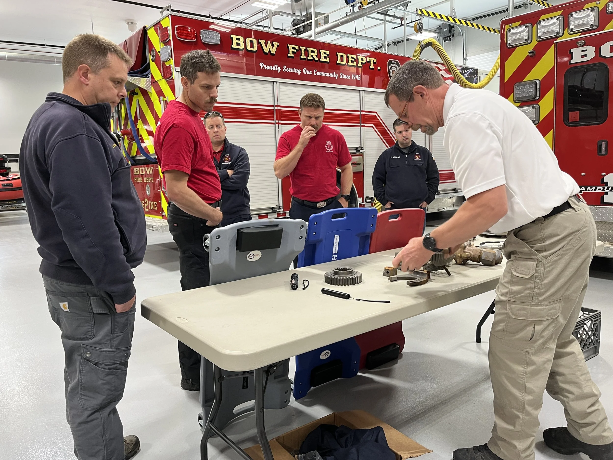 Group of firefighters and instructors gathered around a table, discussing a mechanical part, in front of a red fire truck labeled Bow Fire Dept.
