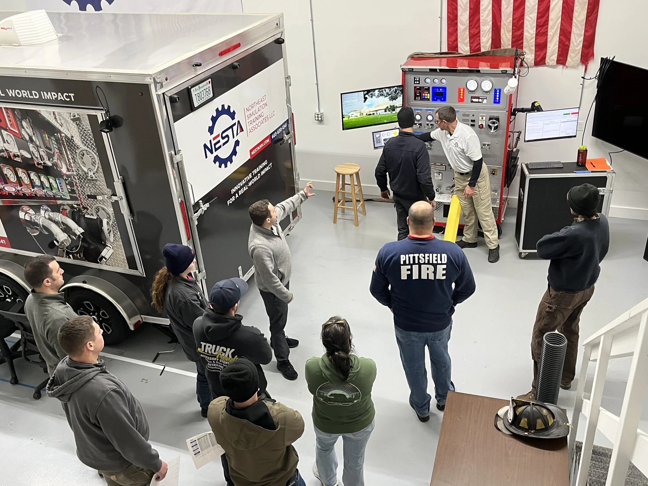 A group of people gathered in a room, listening to a presentation or demonstration involving a large red and silver training simulator or vehicle mockup, with monitors and control panels. An American flag is hung on the wall.