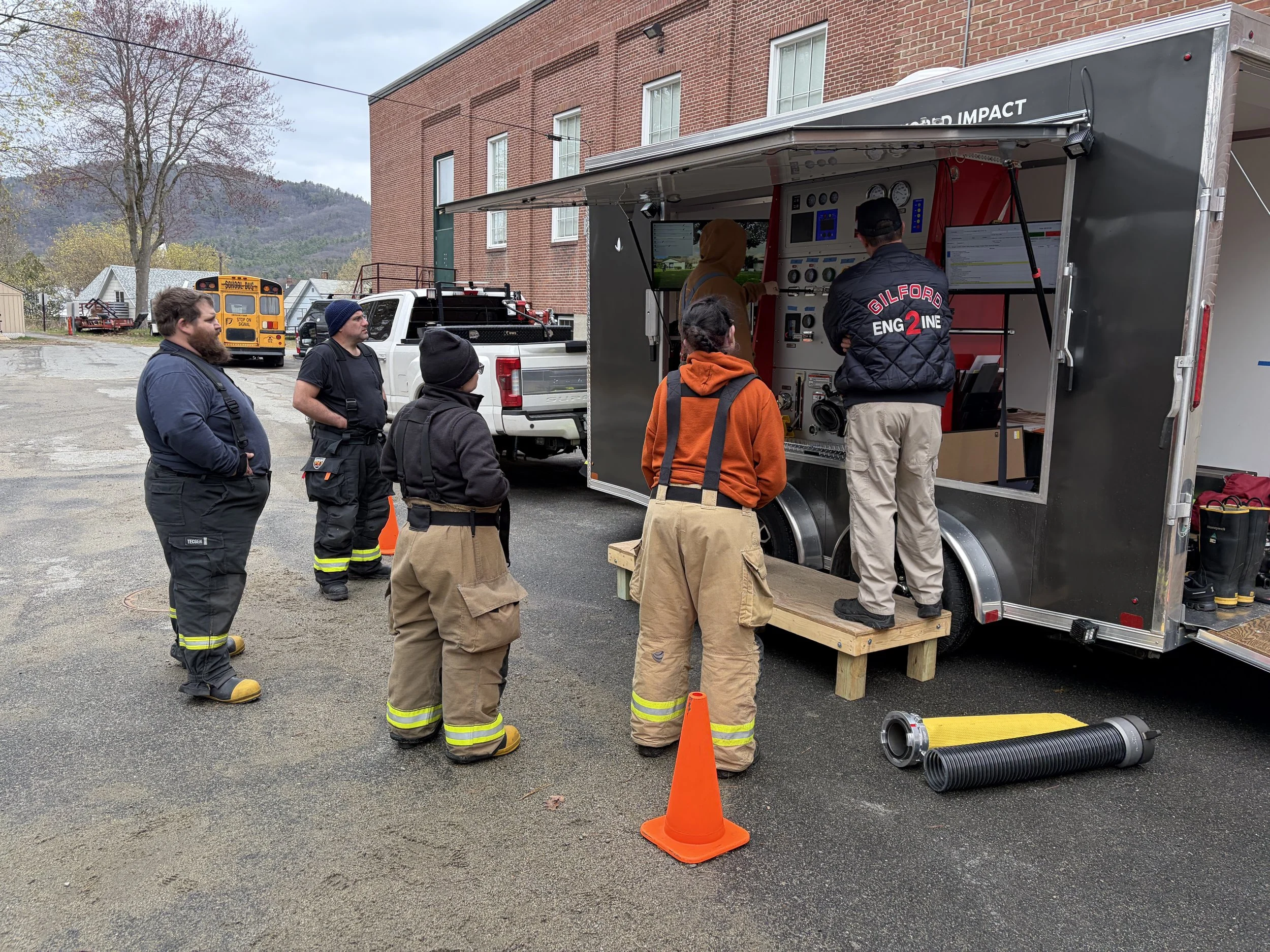 Group of firefighters and instructors gathered around a mobile training trailer with control panels, screens, and gauges outdoors, with a school bus and brick building nearby.
