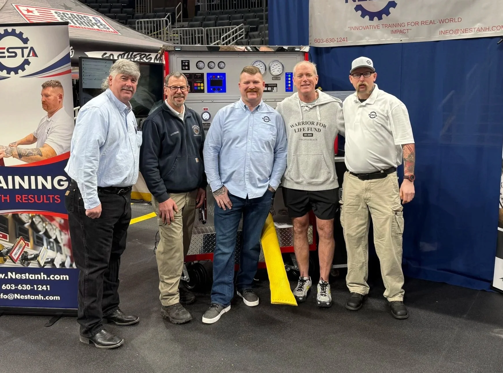 Group of five men standing in front of a red firefighting equipment trailer at an indoor event. They are smiling and dressed casually, with some wearing branded shirts and jackets. Behind them, there are banners and seating in the background.