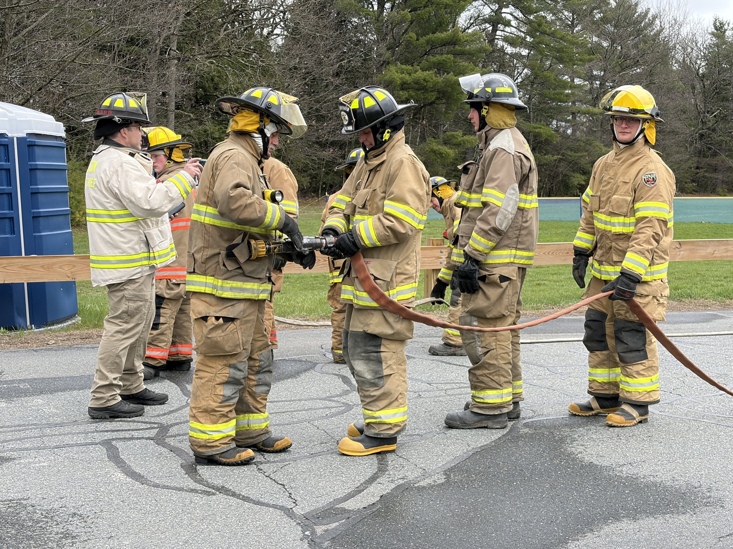 Group of firefighters in tan uniforms and yellow helmets standing on a cracked roadway, with some wearing gloves and holding tools, surrounded by trees.