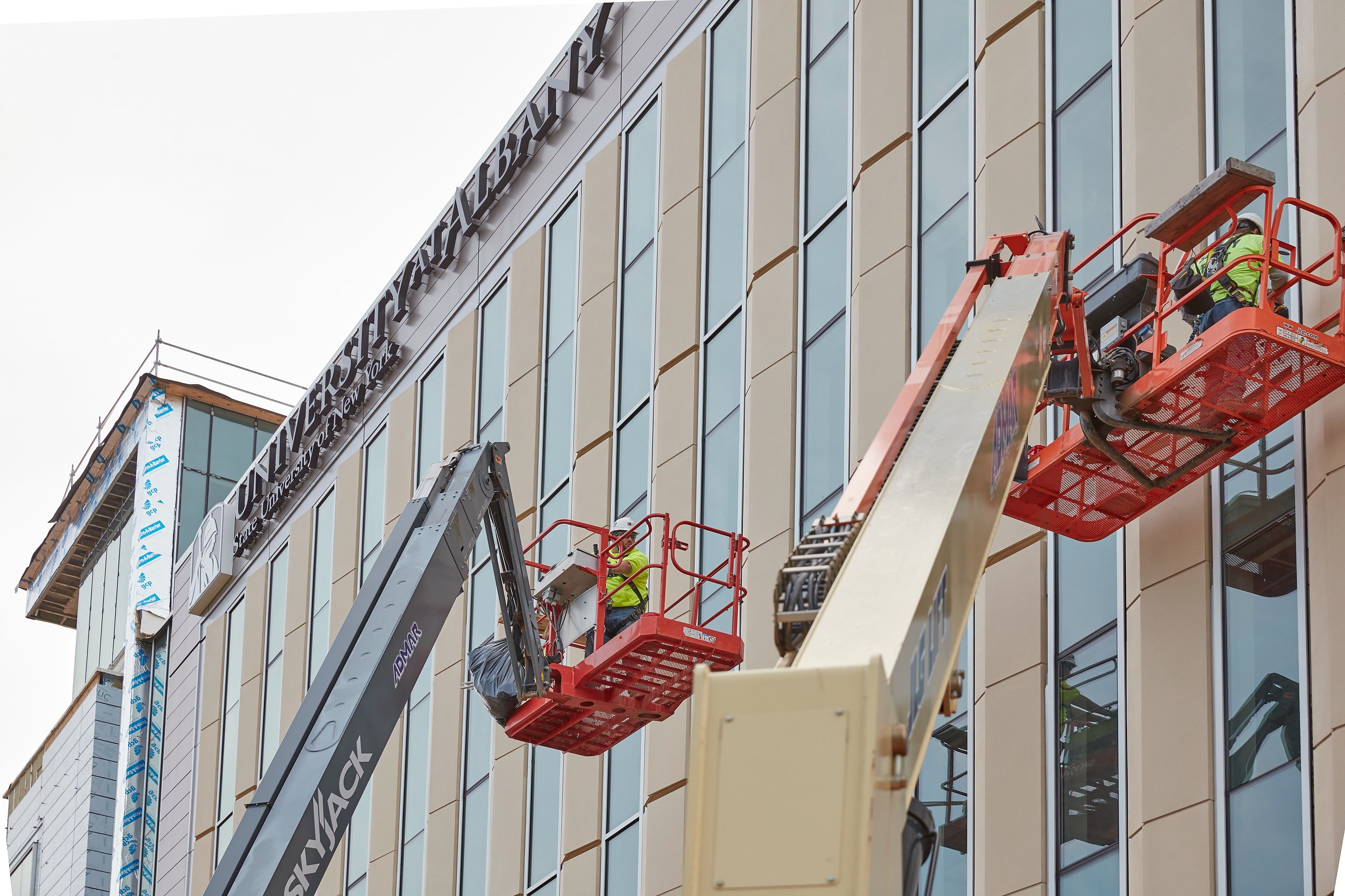 Three construction workers in safety gear operate from cherry pickers, working on the exterior of a modern multi-story building with large windows and beige panels, capturing a moment of expert jobsite photography.