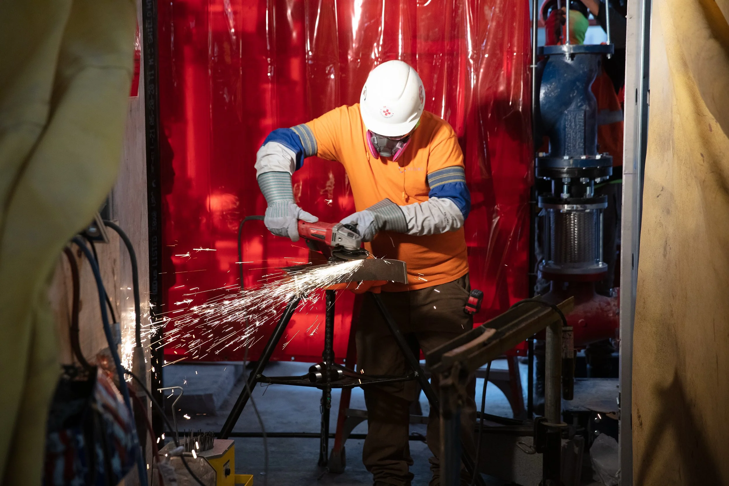 A construction worker in safety gear uses a power grinder, creating sparks. Captured as part of detailed jobsite photography, the worker is positioned in front of a red curtain and surrounded by equipment.