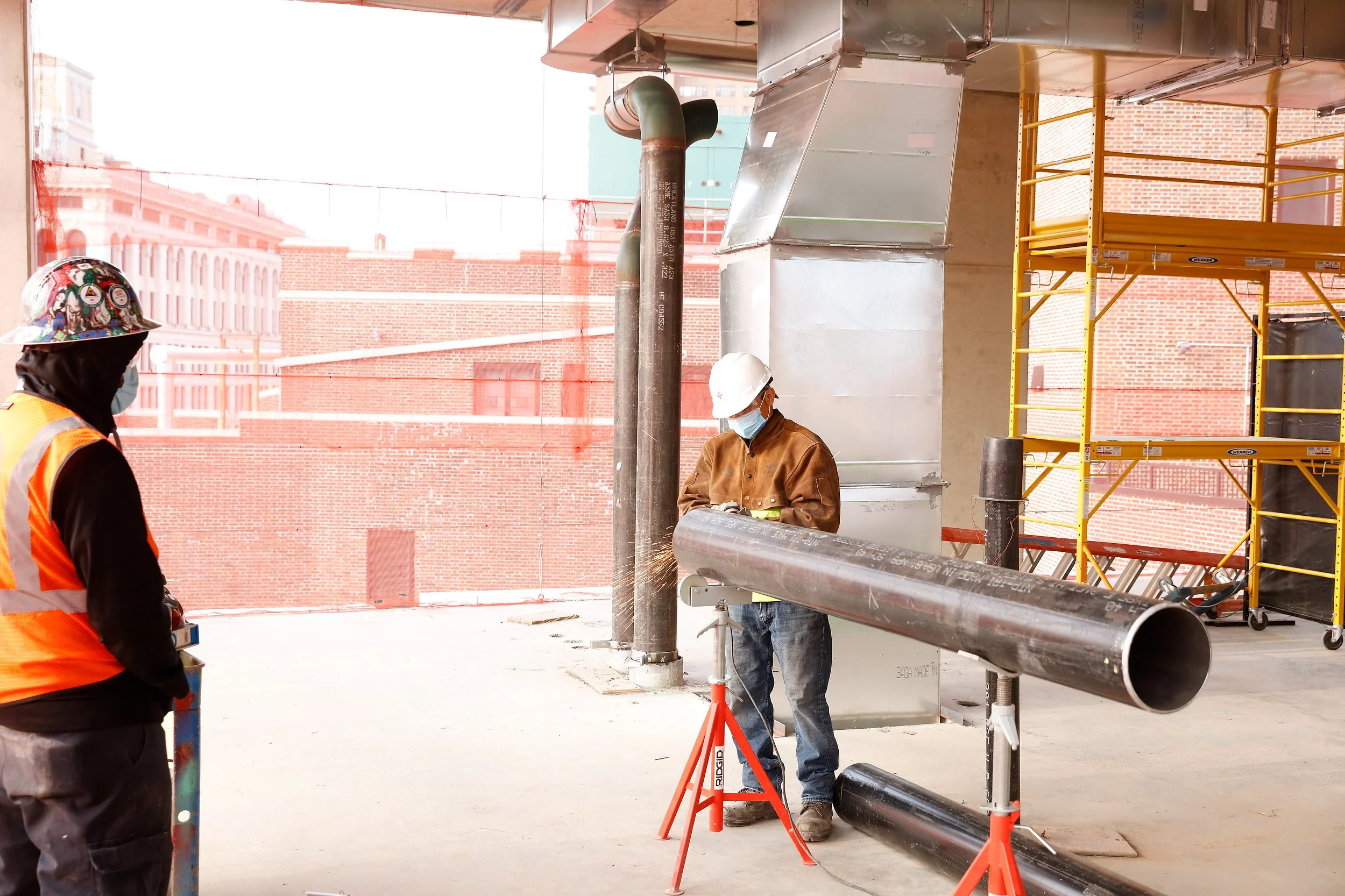 Two construction workers in safety gear work indoors, captured in a moment of jobsite photography. One worker stands near a large metal pipe supported by stands, while the other observes. Scaffolding and buildings are visible in the background.