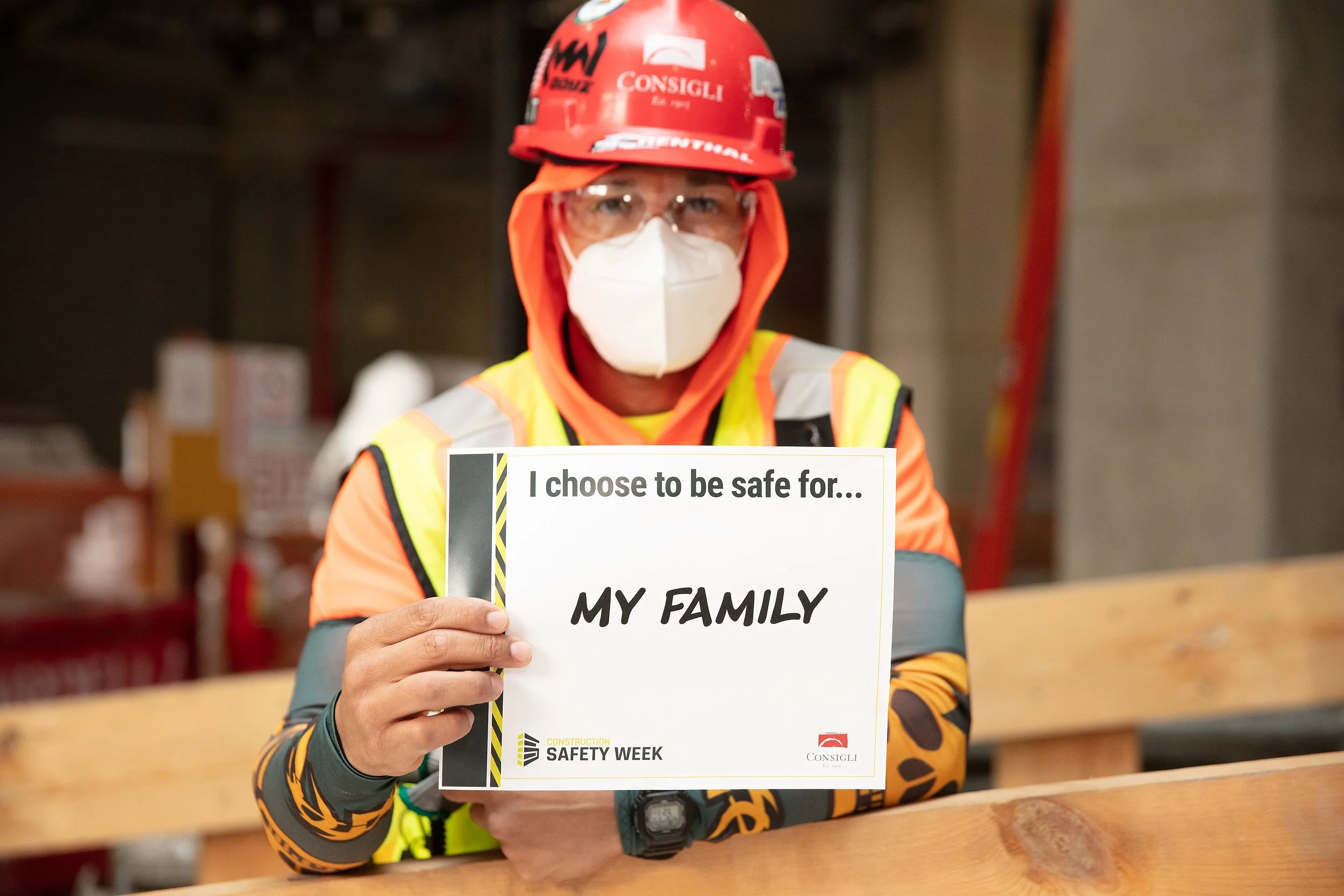 Construction worker in safety gear and a mask holds a sign that reads, “I choose to be safe for... MY FAMILY.” The background shows a construction site, capturing a powerful moment of construction photography that emphasizes safety and responsibility