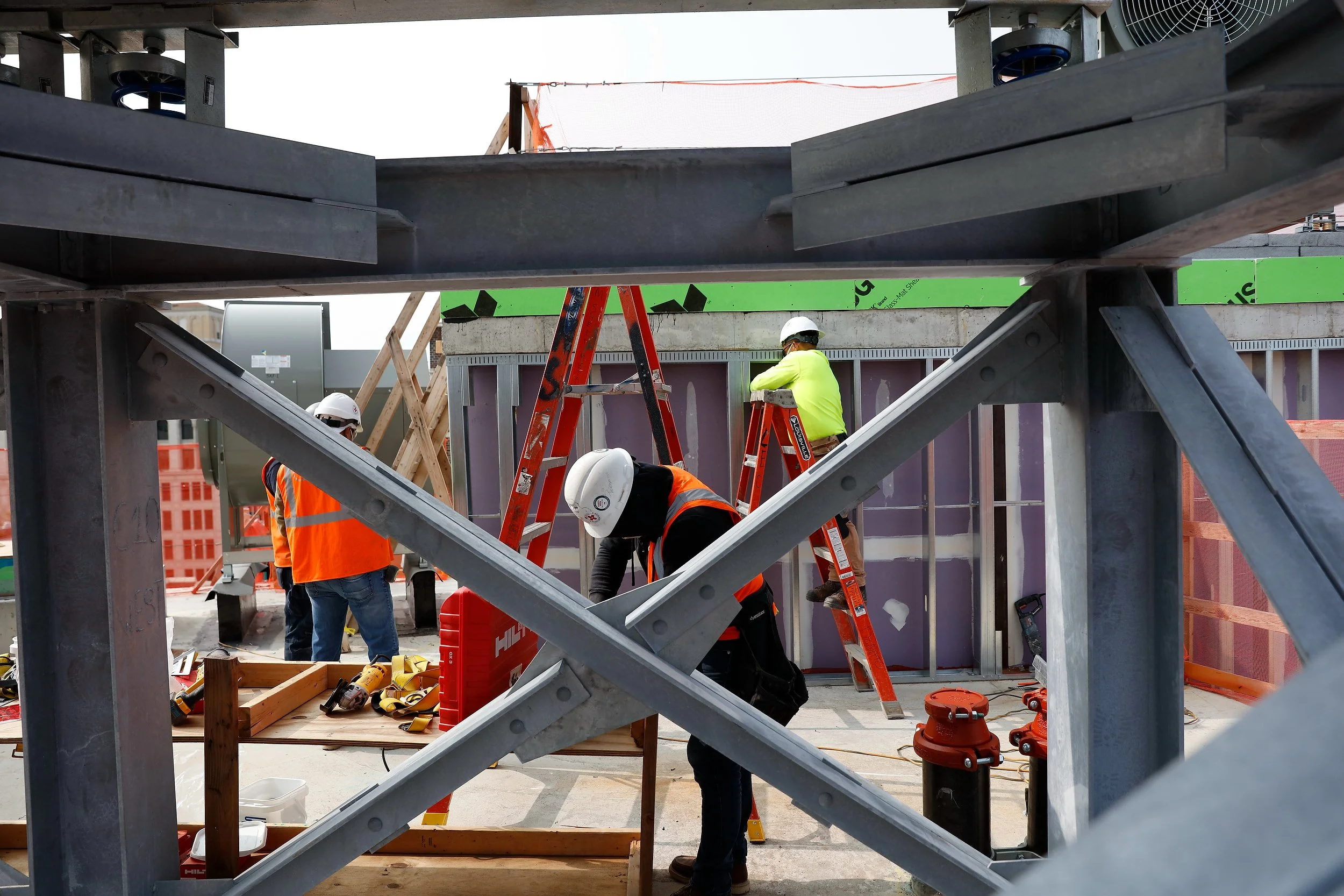 Construction workers in safety gear work around ladders and tools on a building site, framed by large steel beams in the foreground, capturing a moment of precise construction photography.