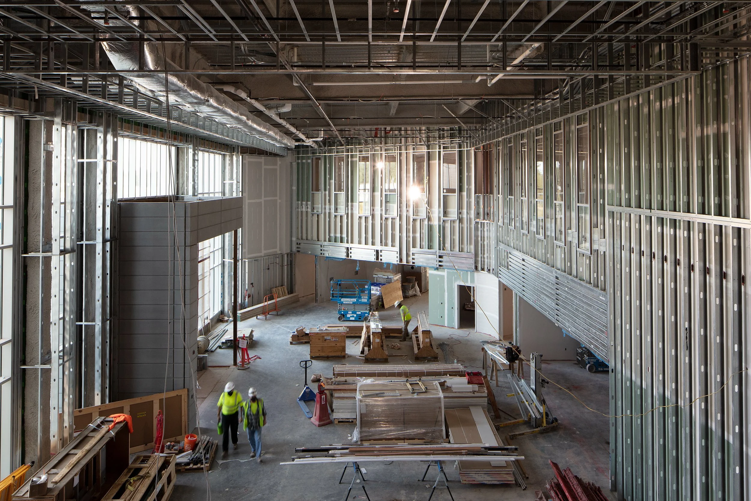 Interior of a large building under construction with exposed metal framing, captured as part of comprehensive construction photography. Several workers in safety vests and helmets are present, along with construction materials and equipment.