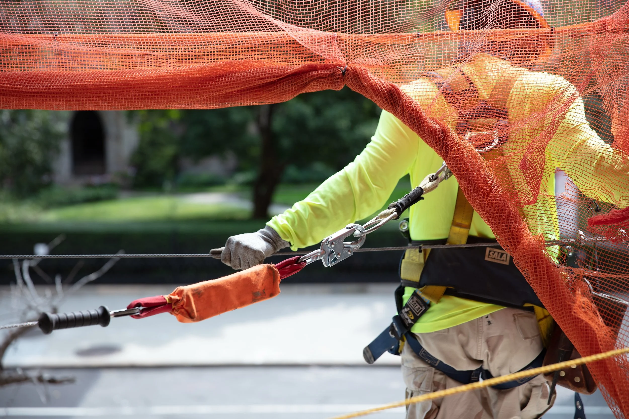 Construction worker in safety gear, tied to a harness, adjusts an orange safety net on a worksite. The worker’s face is not visible; focus is on hands and safety equipment, providing detailed jobsite photography for accurate documentation 