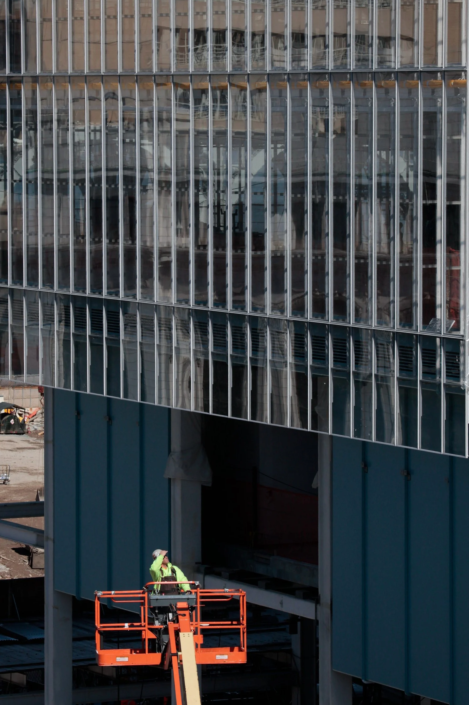 Construction worker in green safety jacket and helmet operating an orange boom lift outside a modern glass building to perform maintenance or construction work.