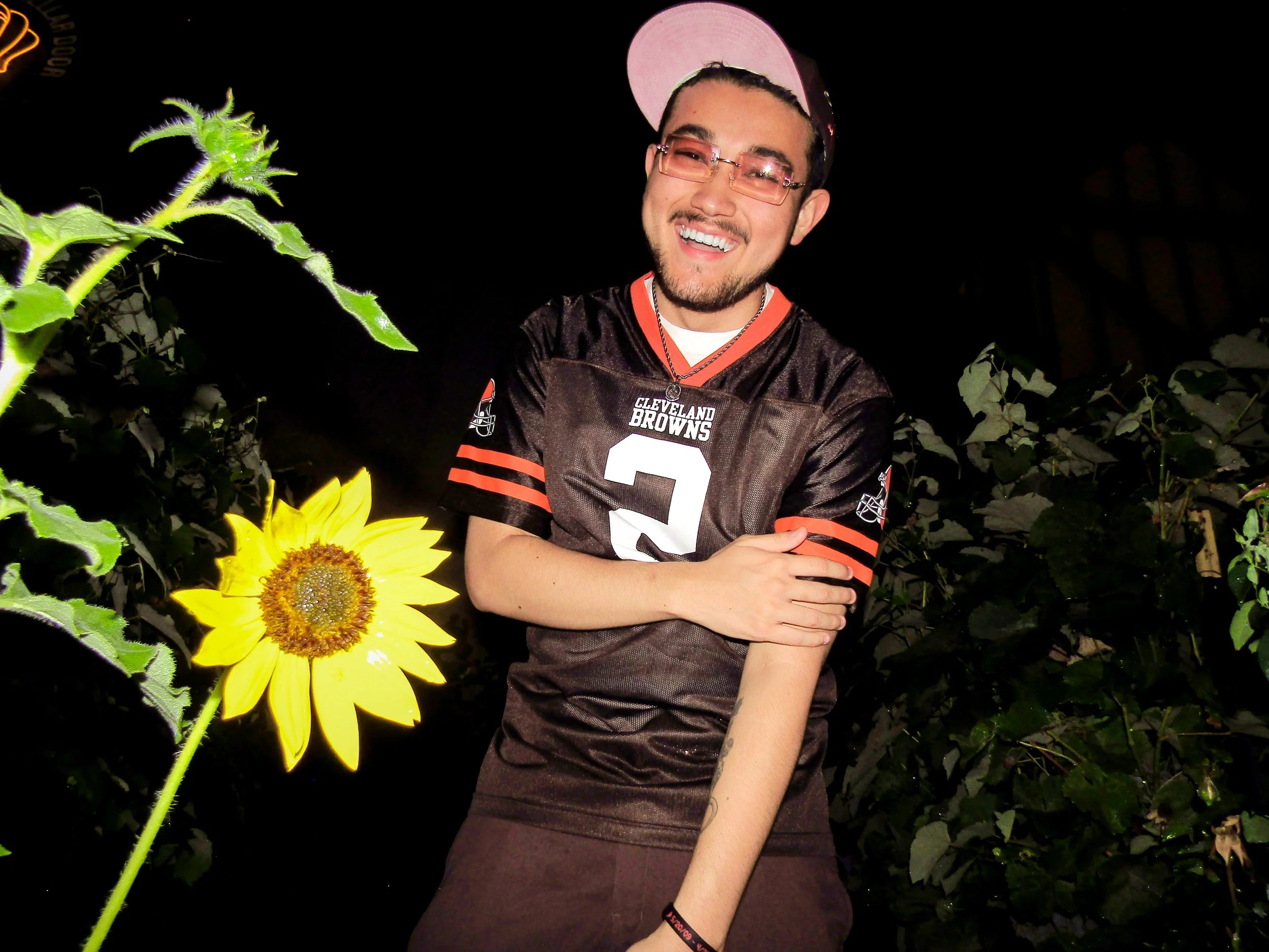 A young man with glasses and a beard wearing a Cleveland Browns football jersey and a pink cap, smiling and standing in front of a sunflower and greenery at night.