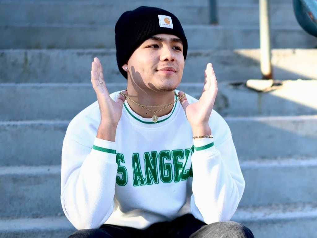 A young man sitting on concrete steps, wearing a black beanie, white Los Angeles sweatshirt, and gold jewelry, with hands raised around his face.