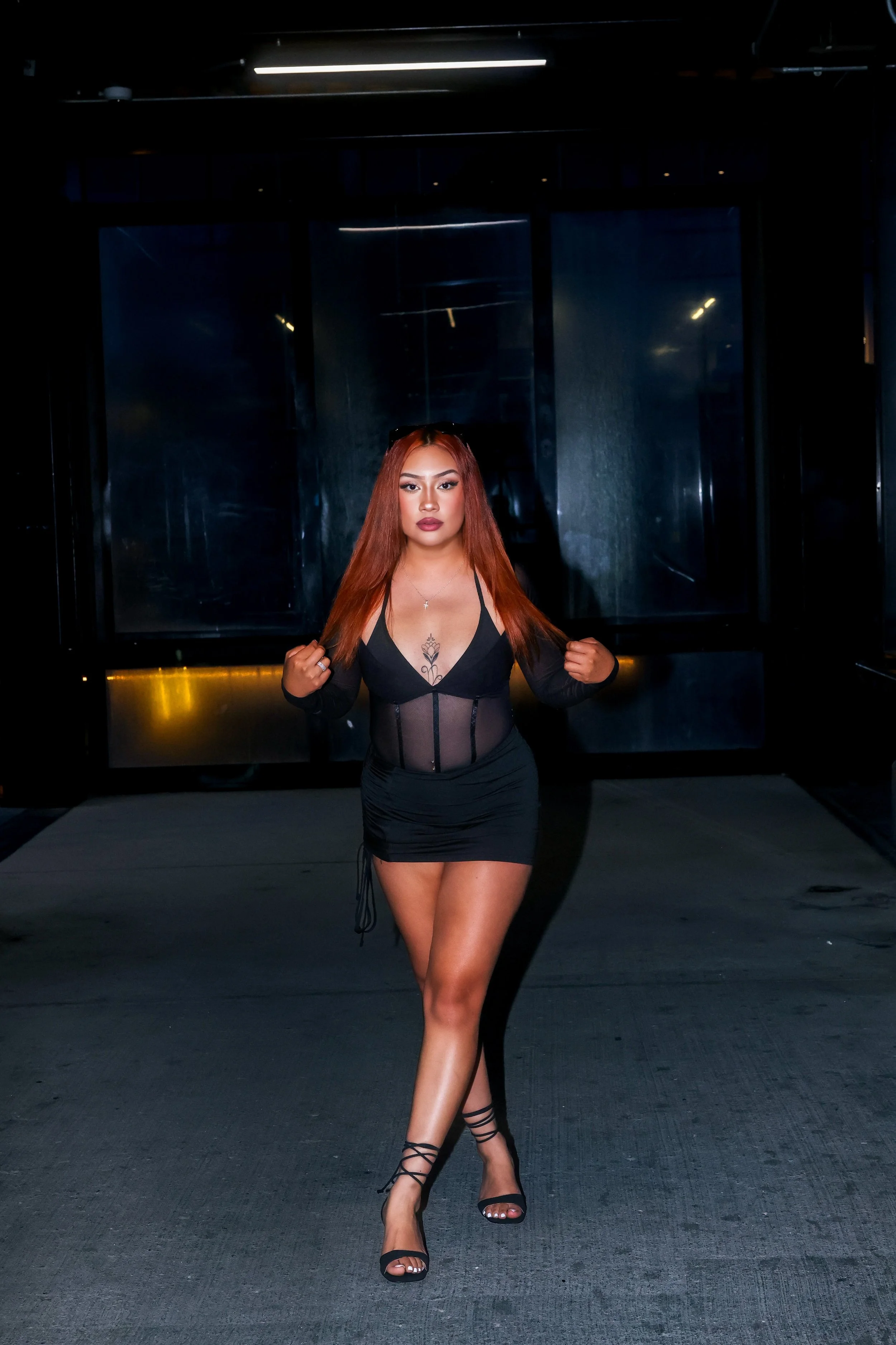 A woman with long orange hair wearing a black sheer dress and high heels walking in a dark indoor setting.
