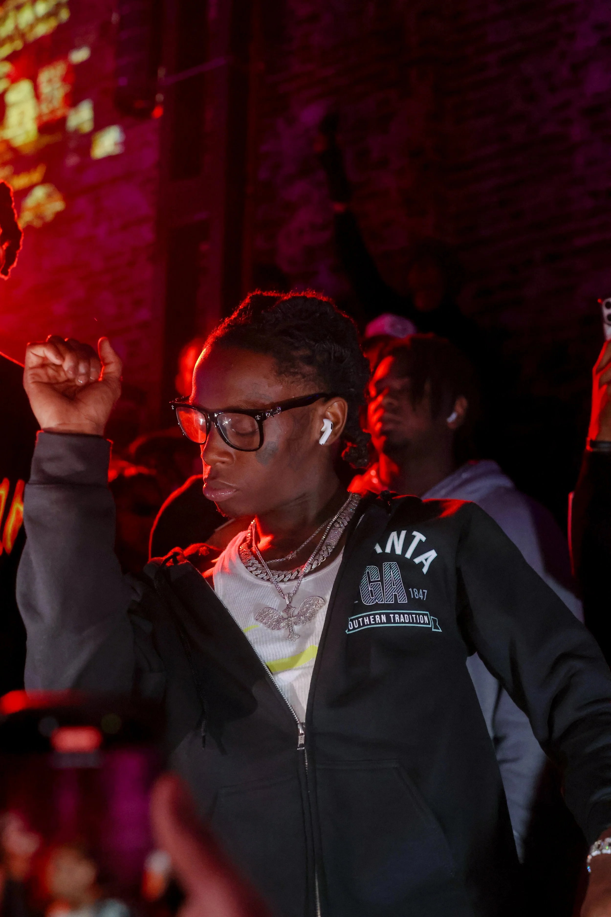 A young person with glasses dancing with a raised fist in a dimly lit room with purple and red neon lights, and other people in the background.