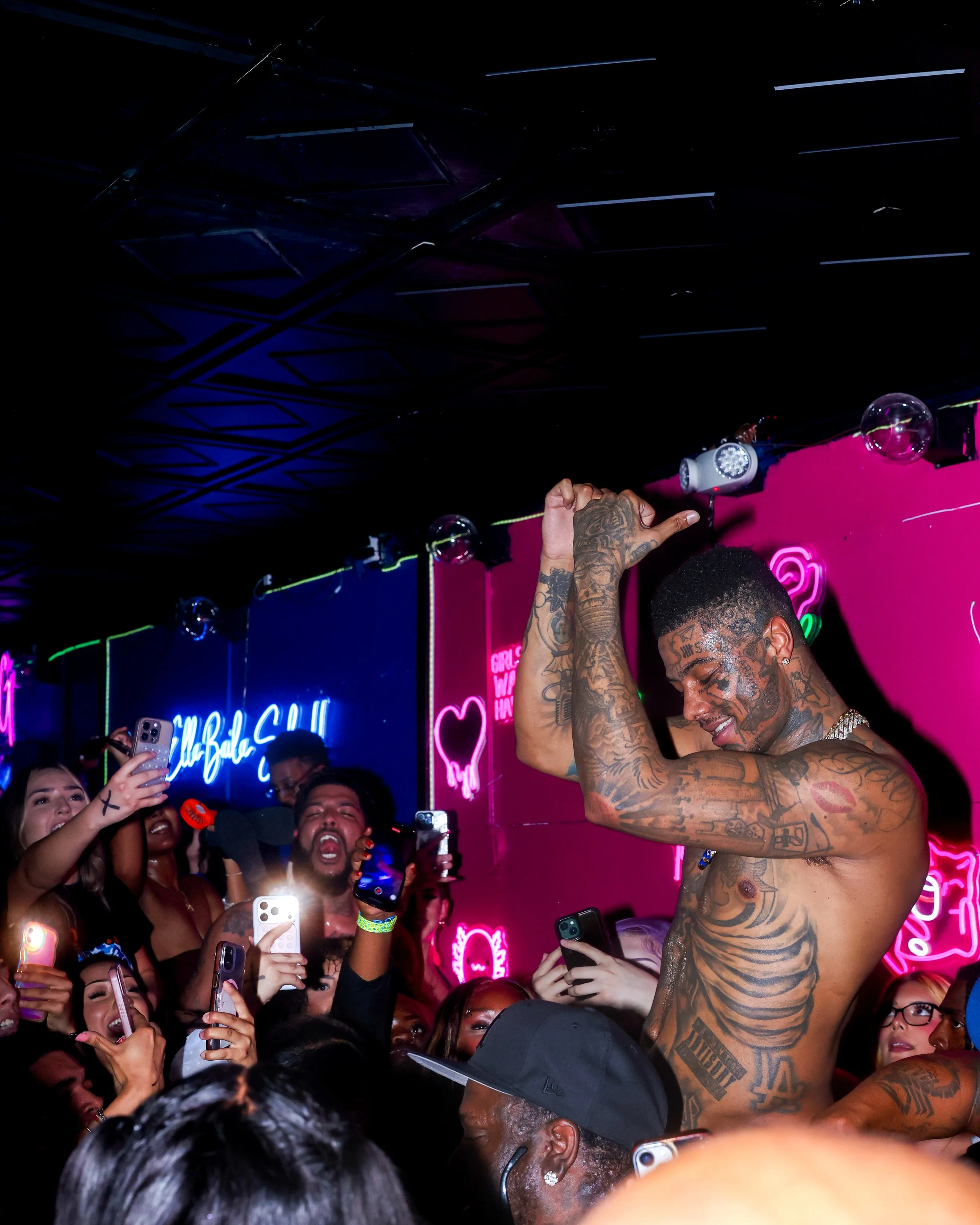 A shirtless man with tattoos dancing or posing amidst a crowd at a club or party, with neon pink and purple signs in the background.