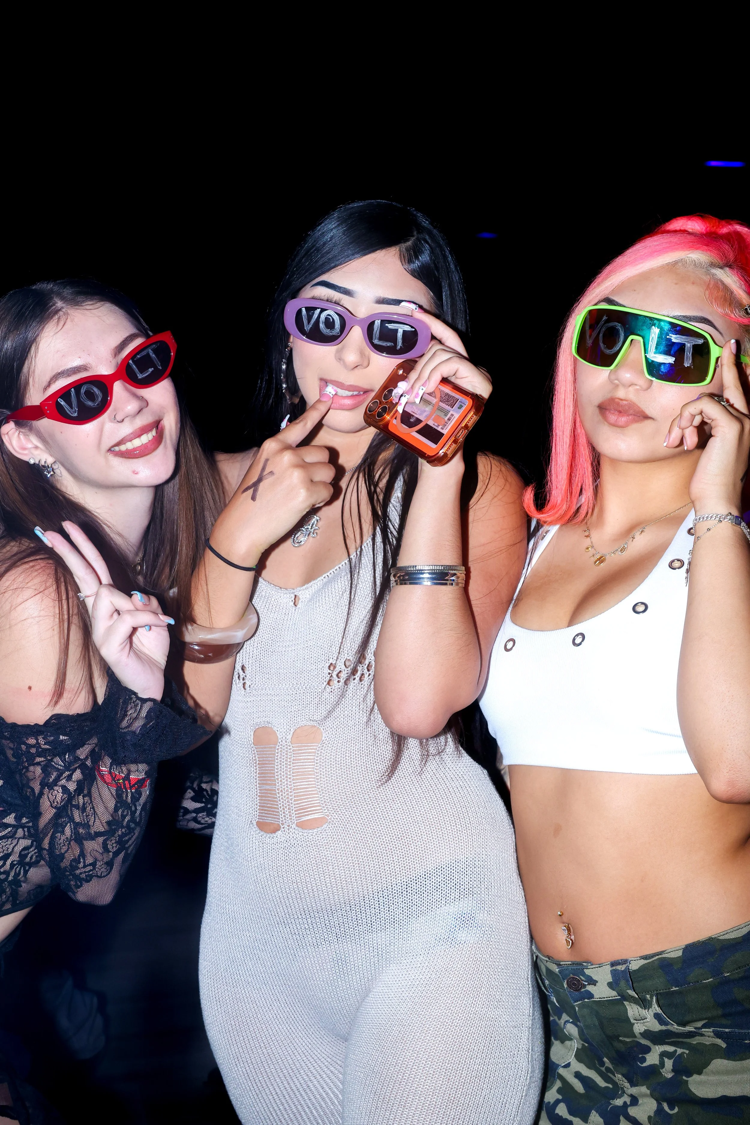 Three women wearing colorful sunglasses with the word "VOLT" on them, smiling, posing together at night.