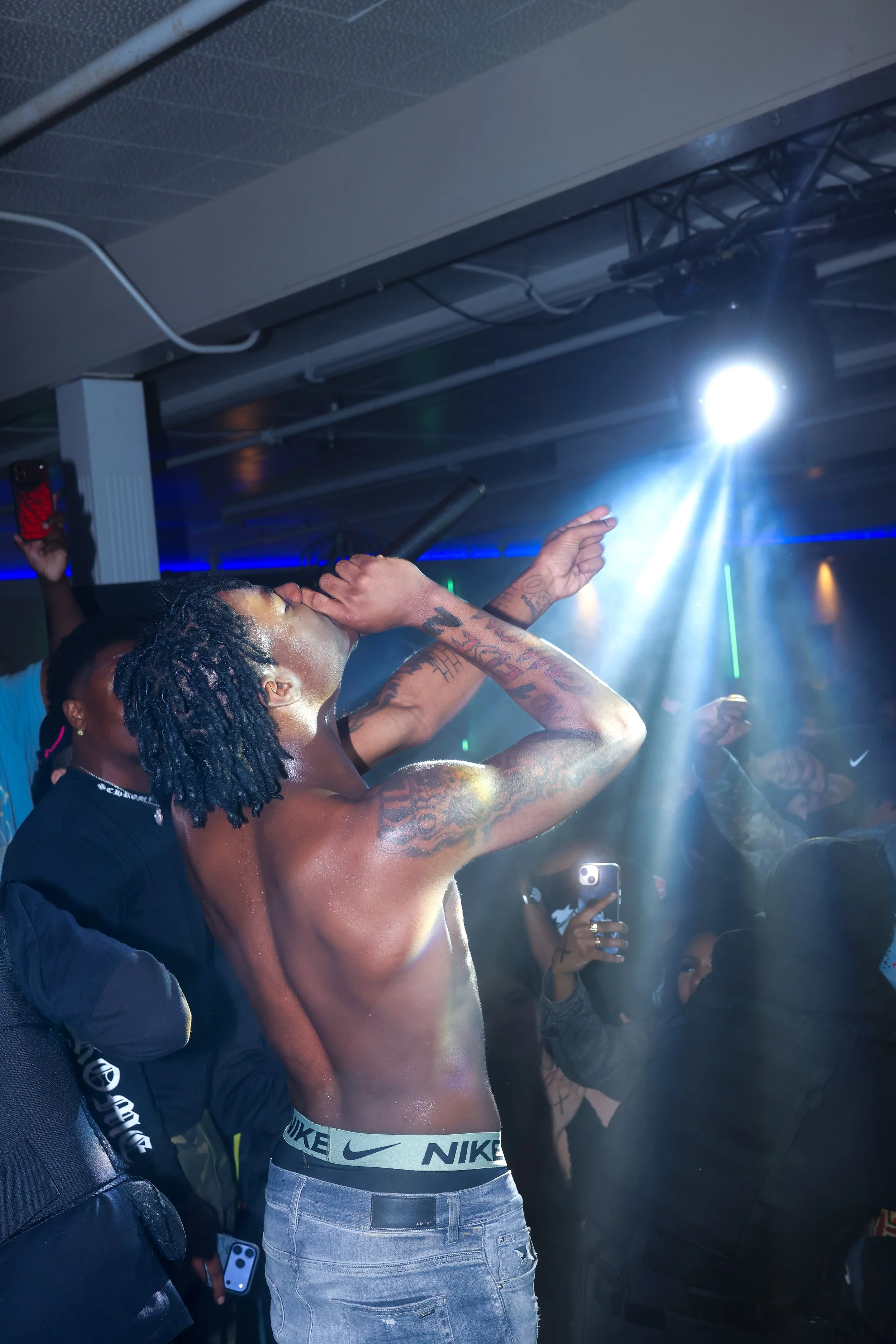 A shirtless man with tattoos and dreadlocks singing into a microphone at a crowded indoor event with blue and white lighting.