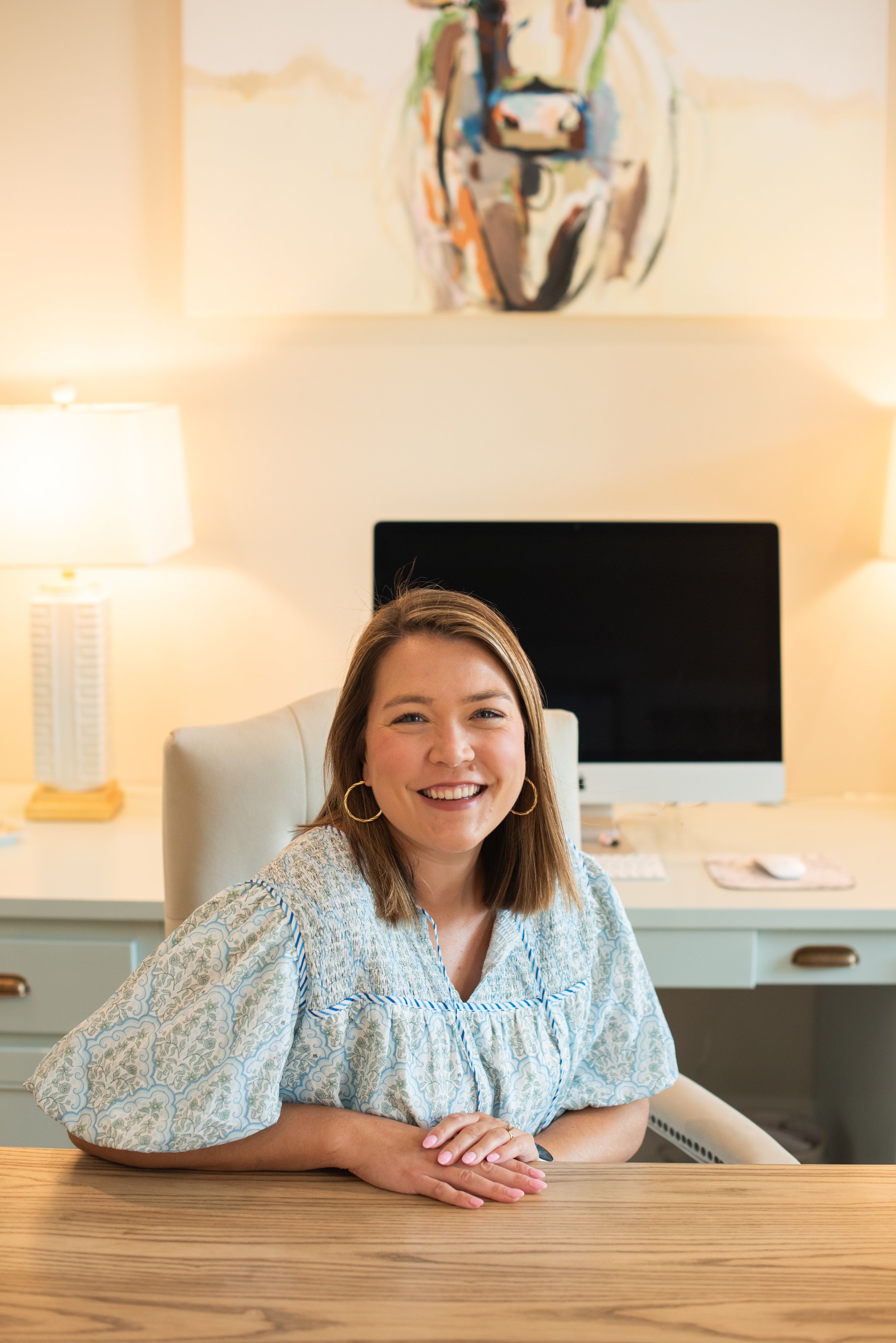A smiling woman with shoulder-length brown hair, wearing a blue and white patterned blouse, sitting at a wooden table in a well-lit room with a large computer monitor behind her, two lamps on either side, and a colorful abstract painting on the wall.