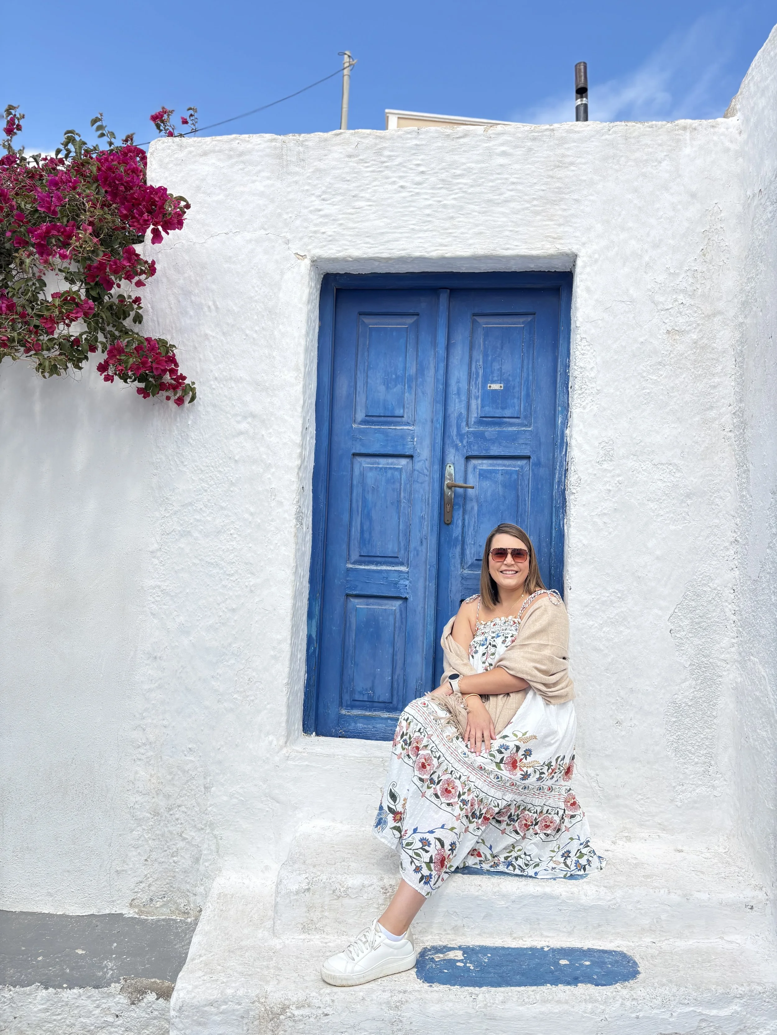 A woman wearing sunglasses, a patterned dress, and a beige shawl is sitting on a white ledge in front of a white stucco wall with a blue wooden door, pink flowers, and a blue sky.