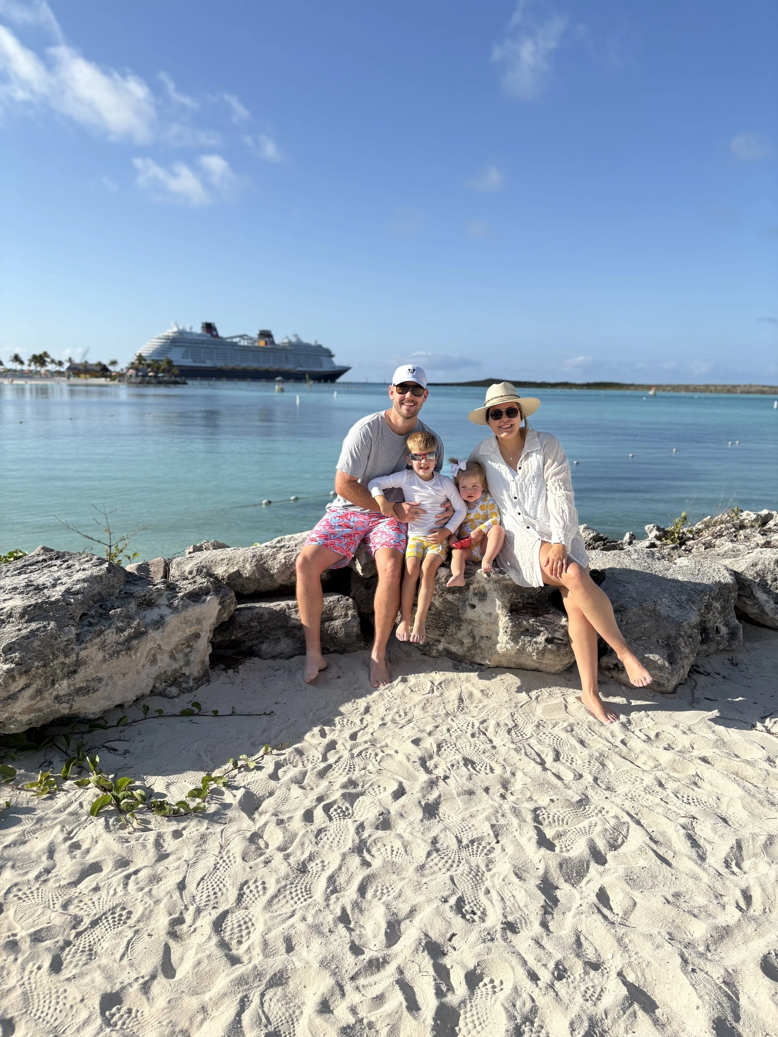 A family of four, including a man, woman, and two children, is sitting and posing on a rocky area at the beach. They are smiling, wearing sunglasses and summer clothing. In the background, there is a large cruise ship on the water and a clear blue sky with some clouds.