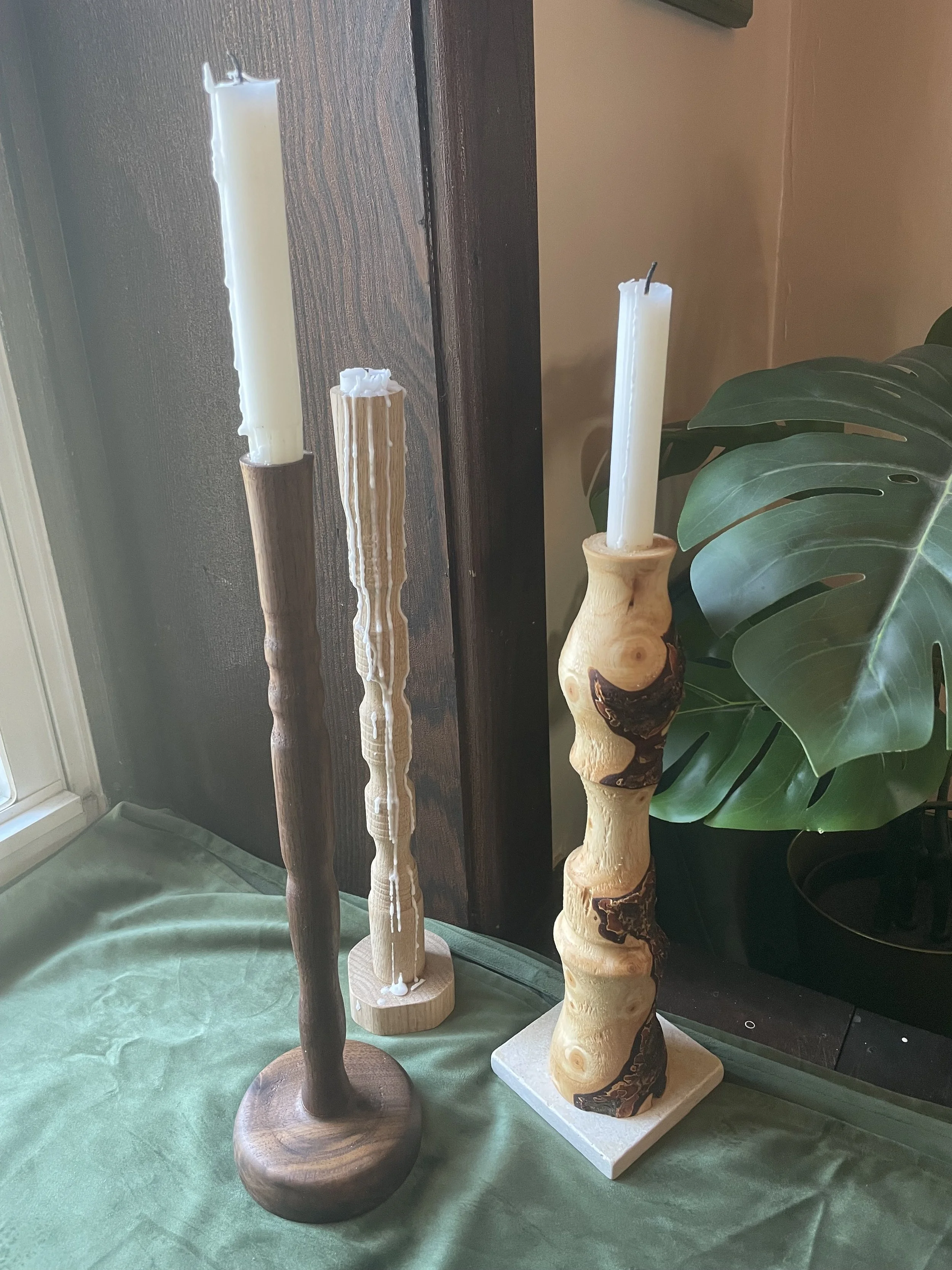 Three tall wooden candles stands on a green cloth surface near a window and a potted plant, with the middle candle being white and the other two darker wood-tone.