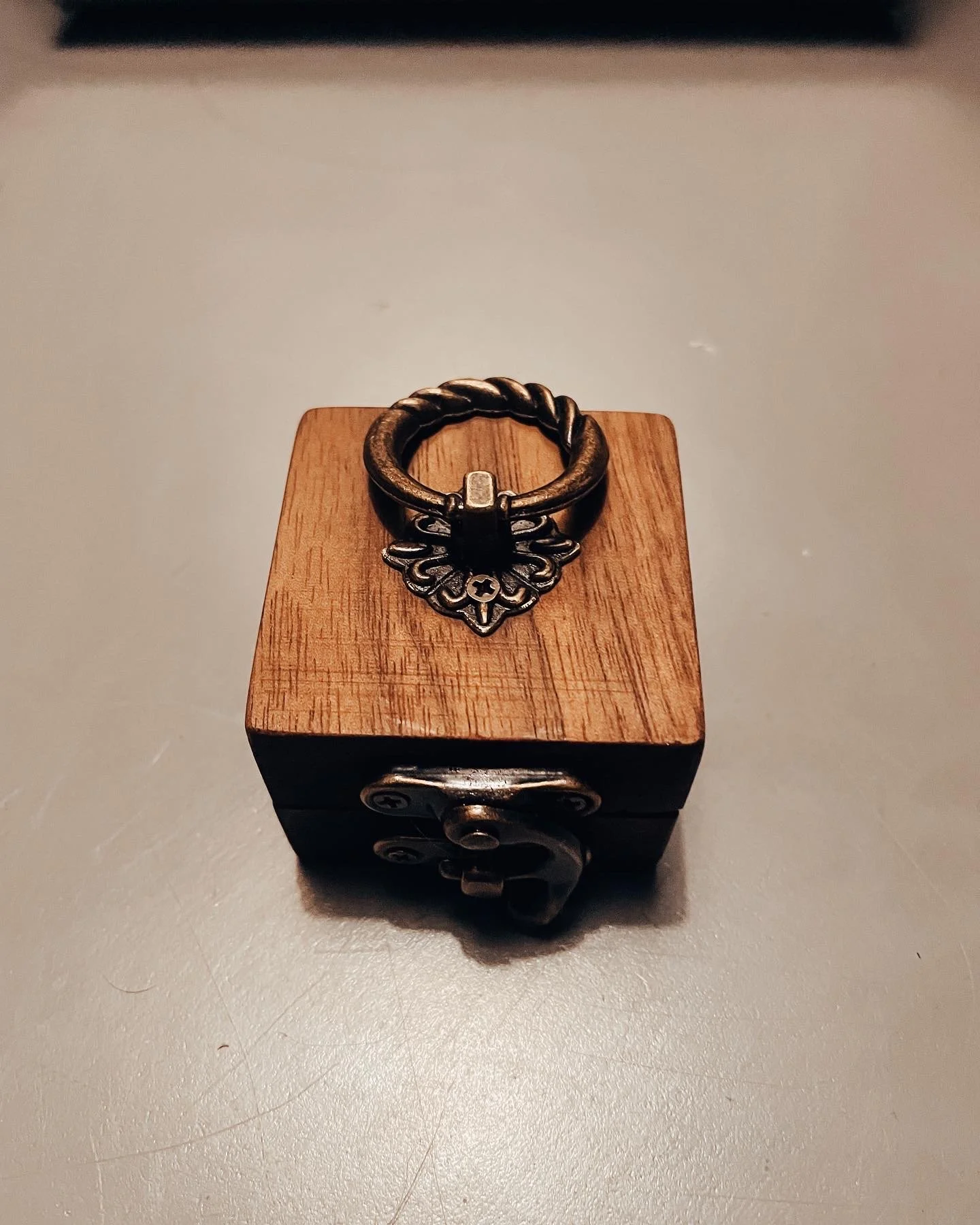 A decorative ring with a textured band and a floral-shaped pendant, placed on a small wooden box on a gray surface.