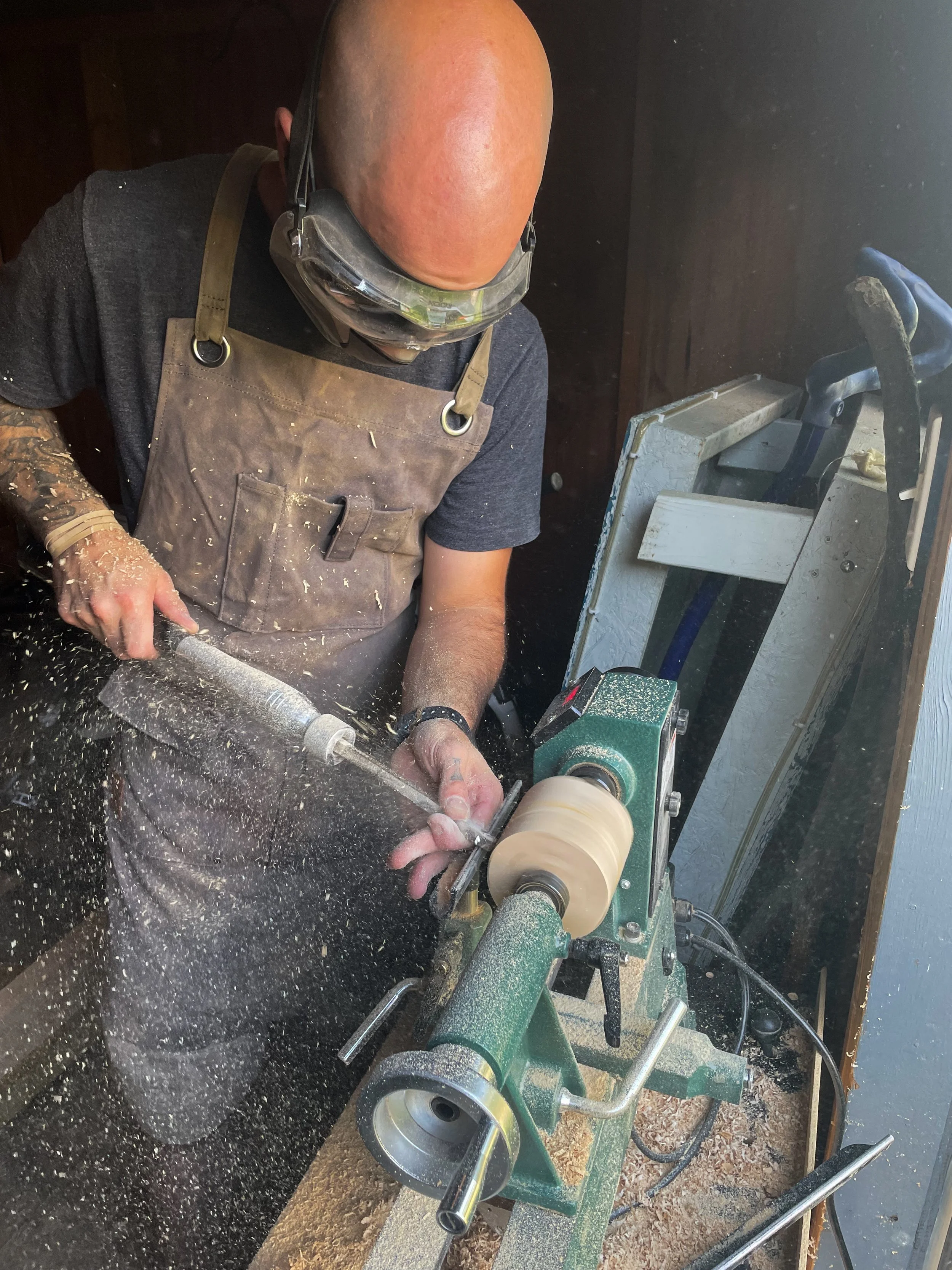 A person using a lathe to shape a piece of wood or material, wearing protective goggles and an apron, with wood shavings flying around.