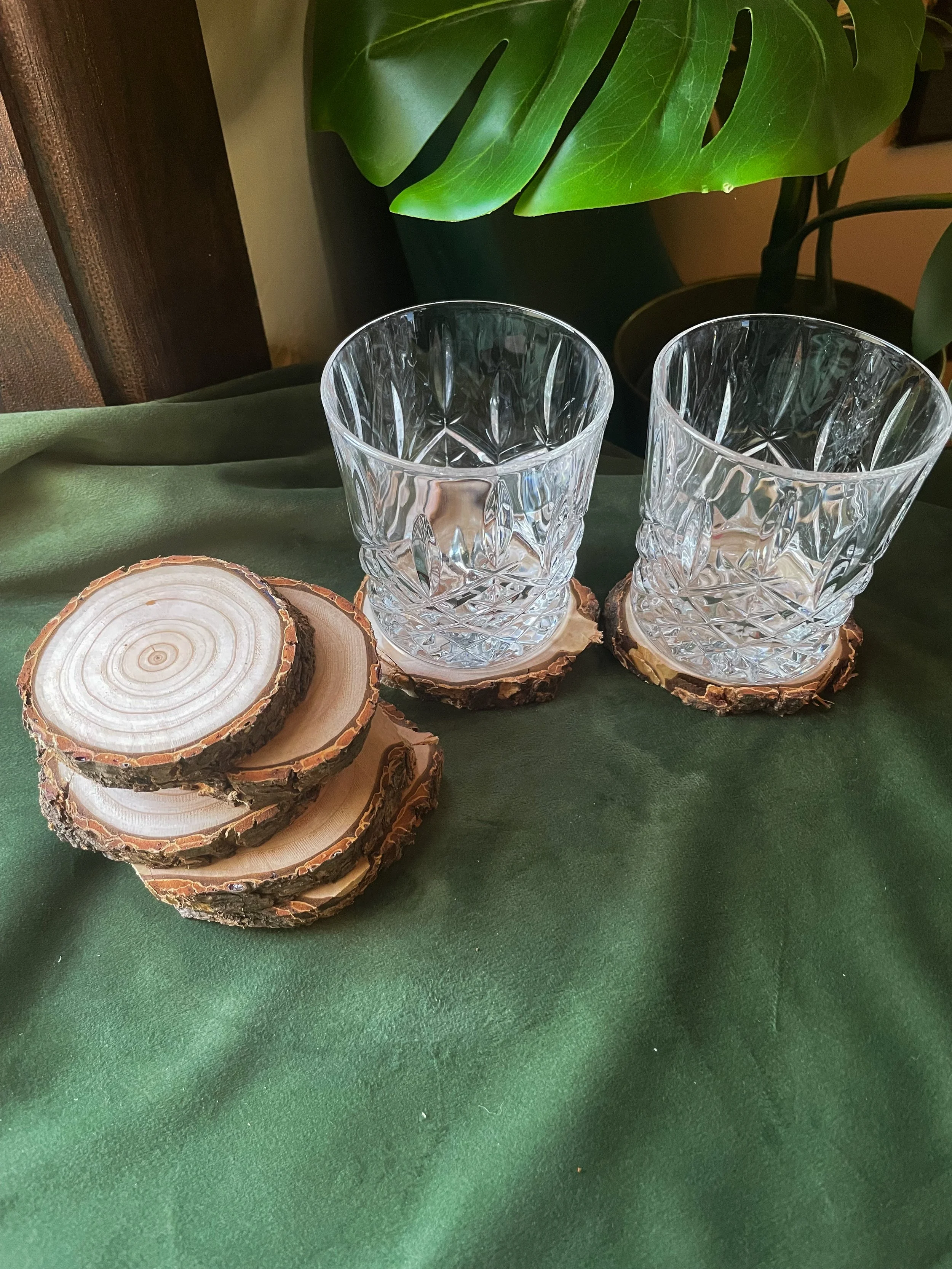 Two clear glass tumblers on wooden coasters, placed on a dark green fabric surface, with large green monstera leaves in the background.