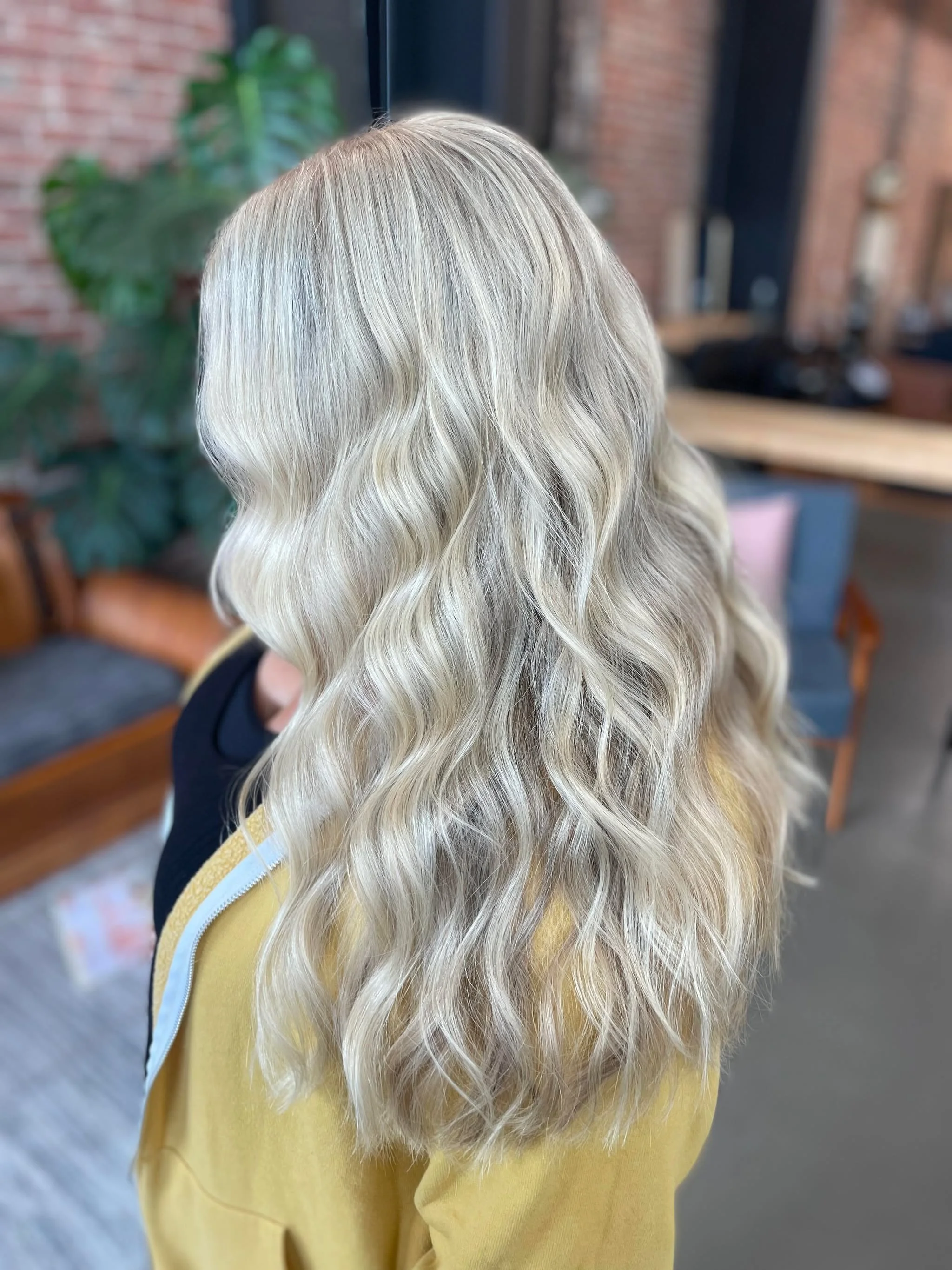 Close-up of a person's head with long, wavy, platinum blonde hair, wearing a yellow sweatshirt. The background shows a plant, brick wall, and indoor furniture.