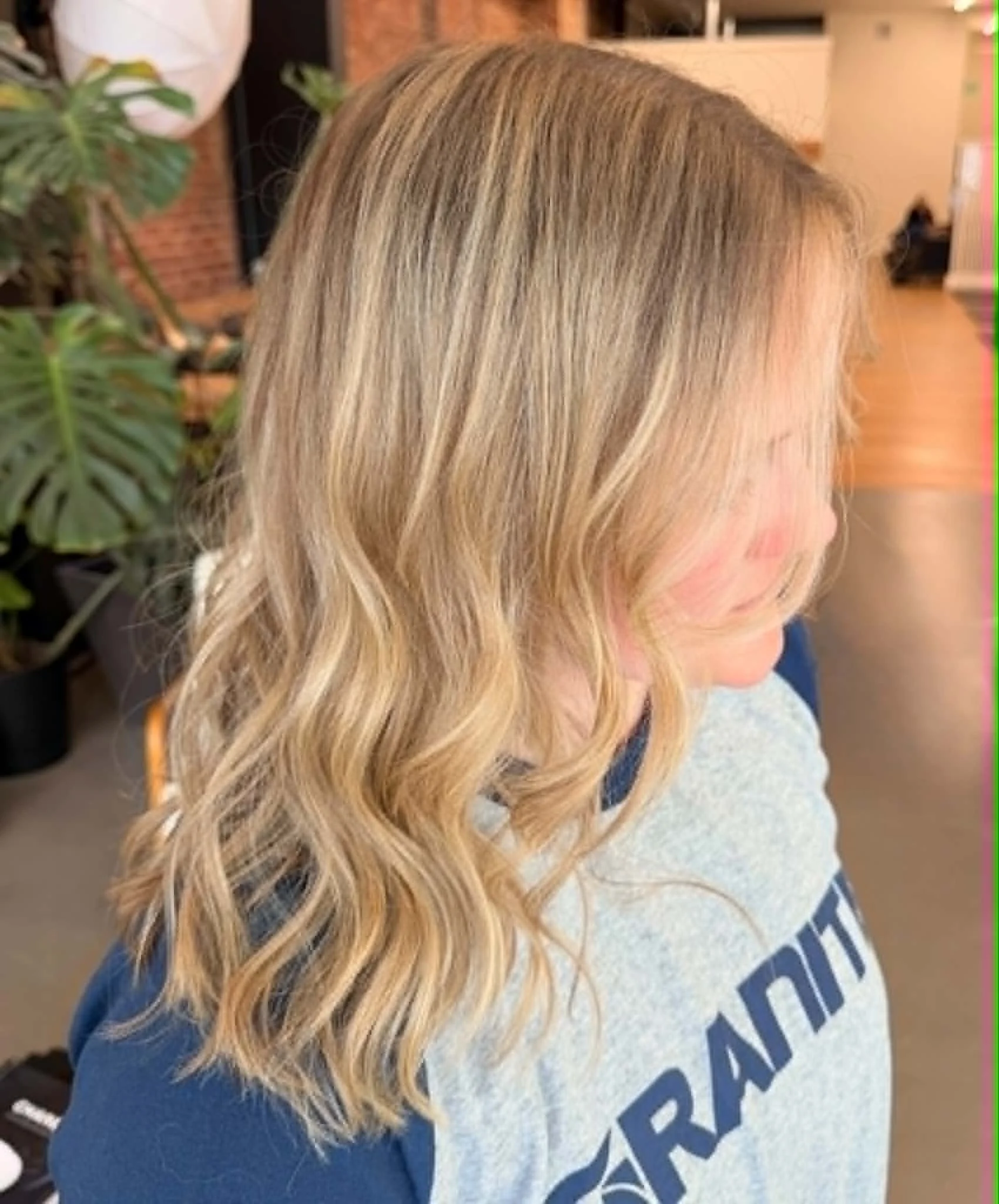Close-up of a woman with long, wavy blonde hair, wearing a gray and blue shirt situated indoors.